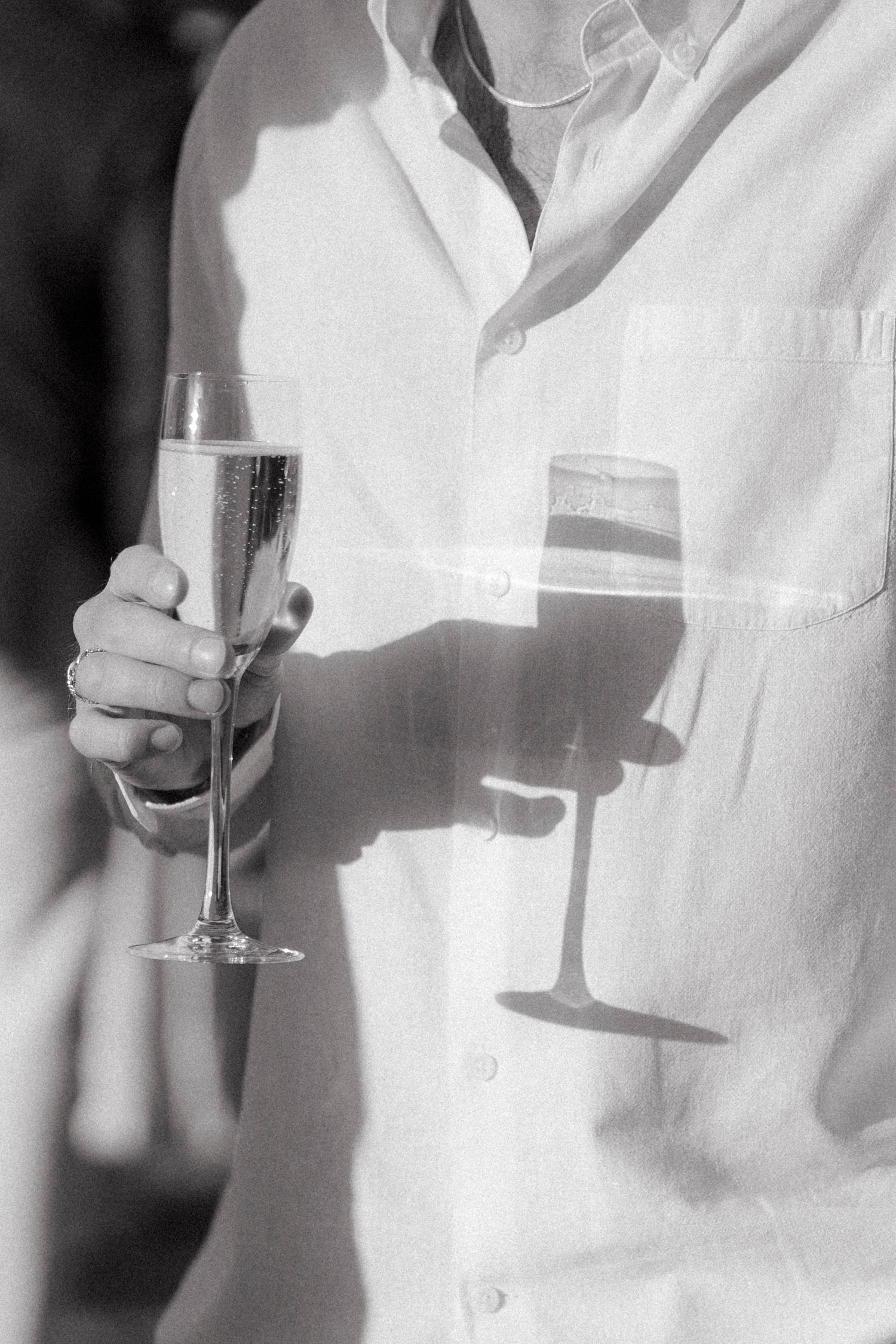 Black and white photo of a man in a linen shirt holding a champagne flute, its shadow cast on his shirt in sunlight.