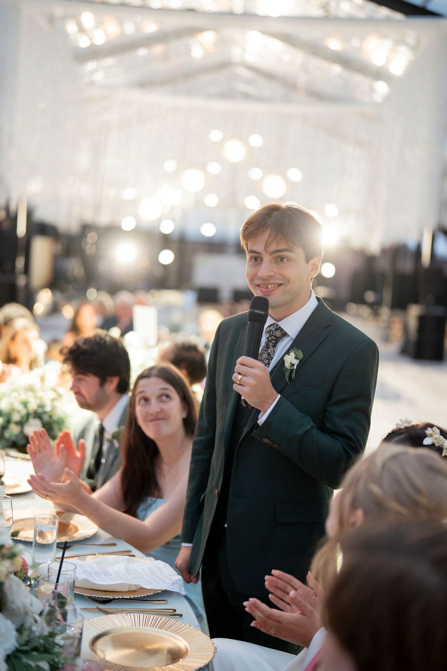 Groom in dark suit gives speech with microphone at elegant wedding reception with string lights overhead.