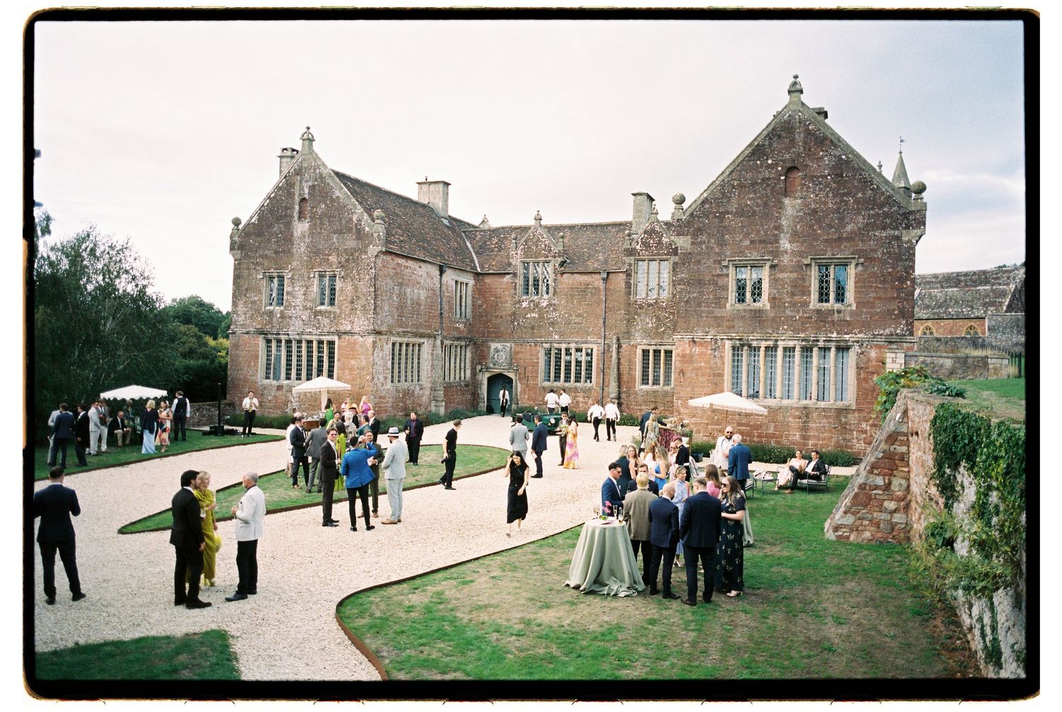 Wedding guests gather outside a historic English manor house venue on a cloudy day.