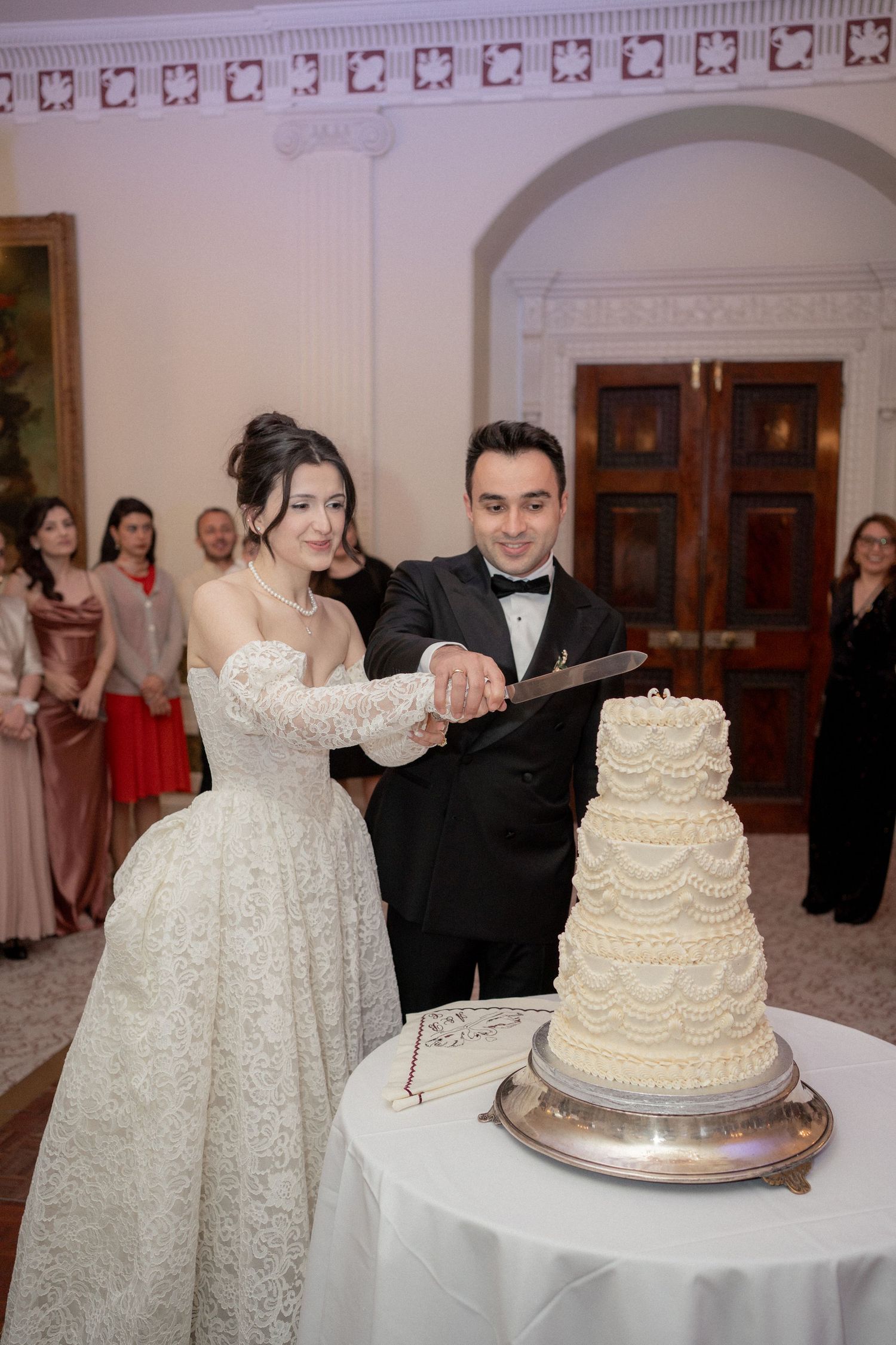Elegant white wedding cake being cut by couple in formal attire at reception venue.