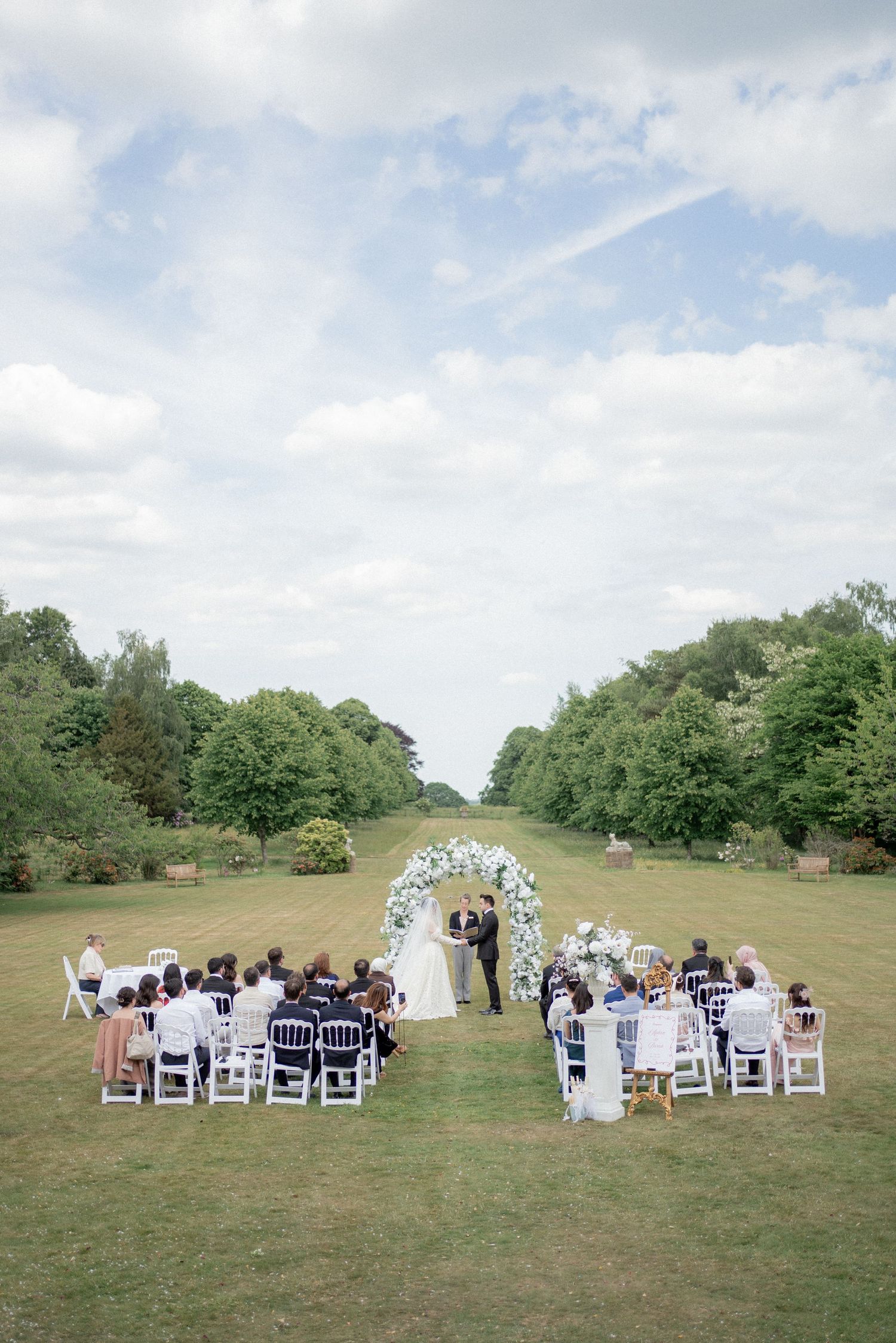 A panoramic view of an outdoor wedding ceremony set up on a grassy hillside with white chairs and natural scenery.