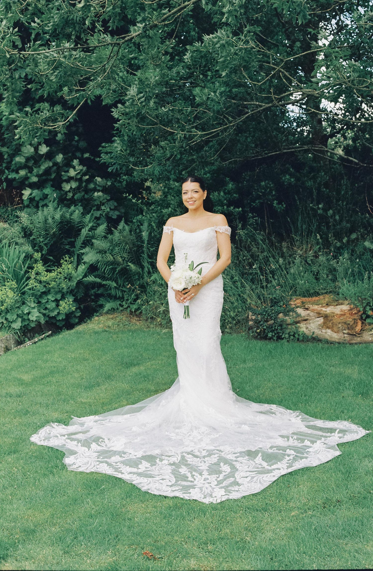 Bride in a white lace wedding gown sitting gracefully on green grass during an outdoor photoshoot.