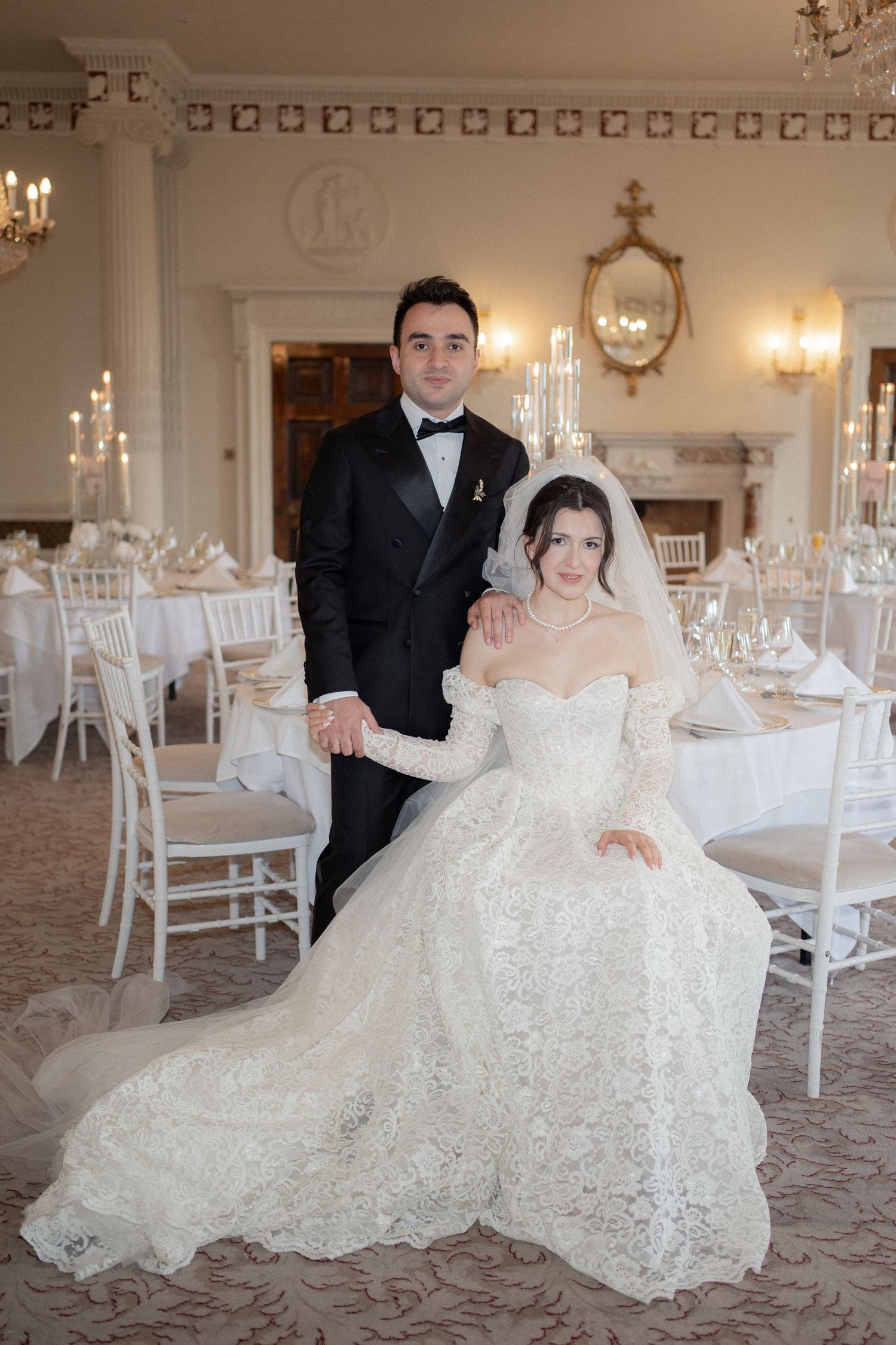 Wedding couple posing in an elegant ballroom with white decor and chandeliers.