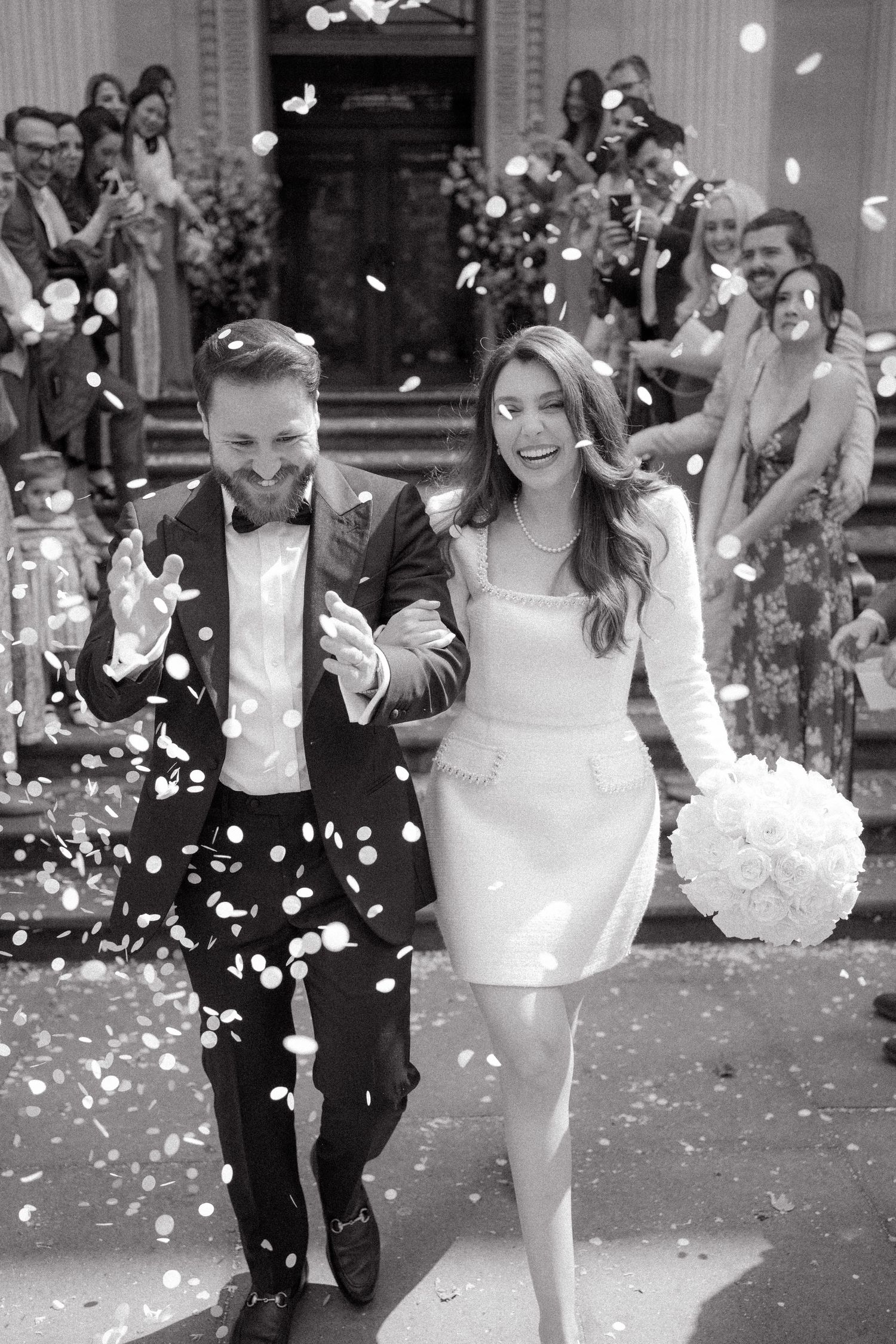 A couple runs through falling white flower petals in formal attire.