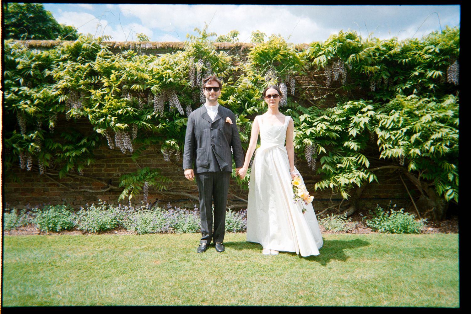 Bride and groom in sunglasses hold hands in front of a brick garden wall covered in climbing vines, on a sunny wedding day.