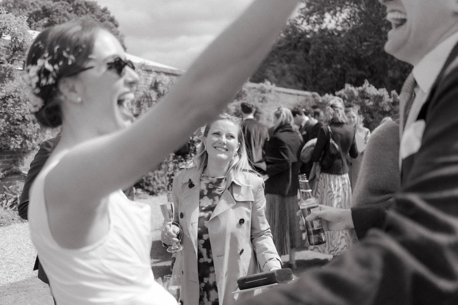 Black and white wedding photo of a laughing bride in sunglasses with her arm raised and a smiling guest holding a drink behind her.