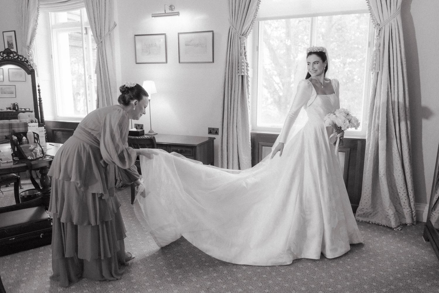 Bridesmaid adjusting the train of a brides elegant white wedding gown in a classic room, black and white photo.