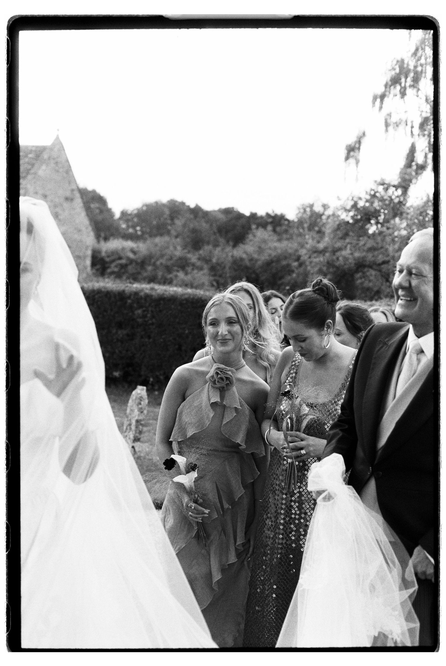 Black and white photo of wedding guests smiling and laughing outdoors, with bride's veil visible in foreground.