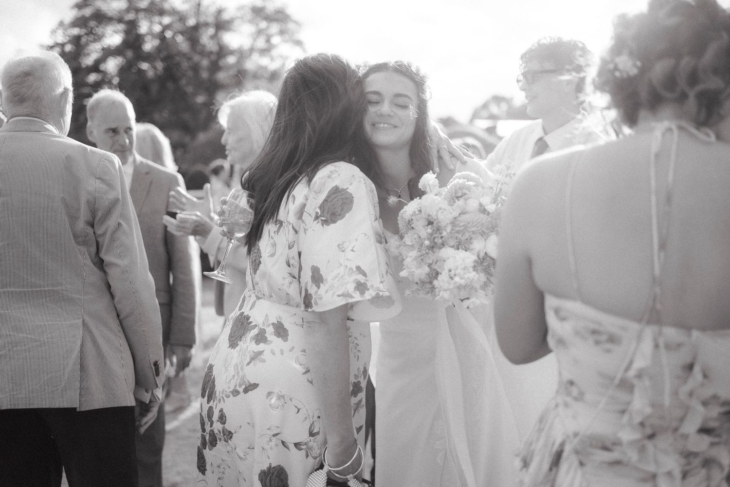 Black and white photo of a bride in a white gown receiving hugs from guests at an outdoor wedding reception.