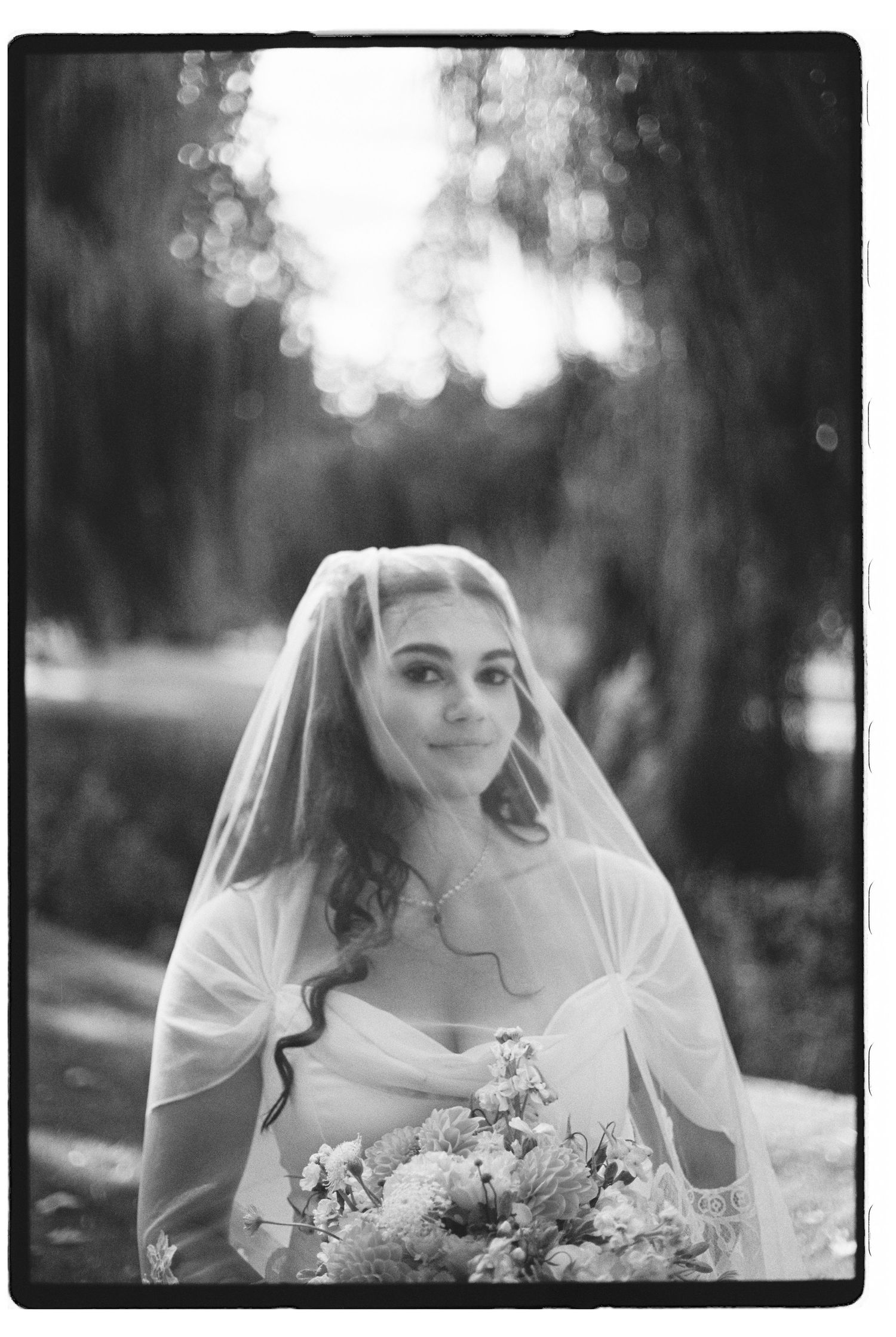 Black and white portrait of a bride wearing a veil and holding a bouquet, smiling outdoors.
