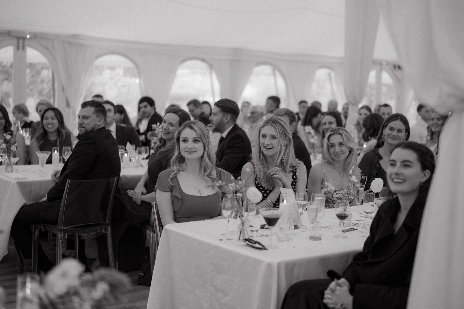 Wedding guests seated at round tables in an elegant white tent reception venue enjoying a formal dinner celebration.
