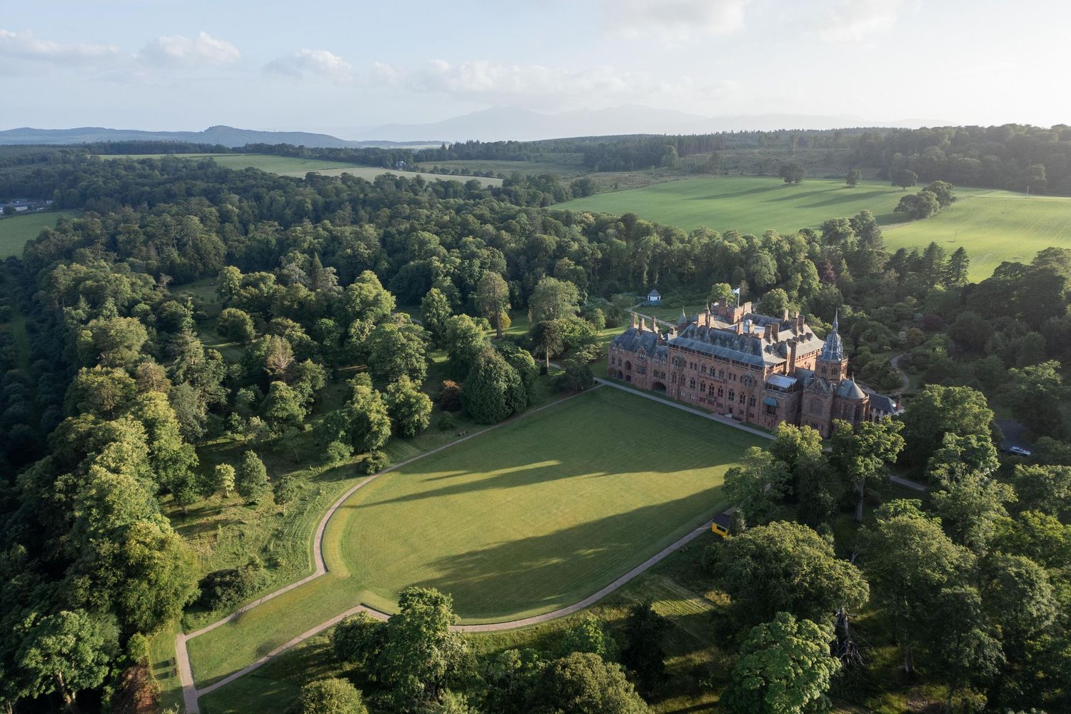 Aerial view of a grand historic mansion surrounded by lush green trees and manicured lawns in a scenic countryside.