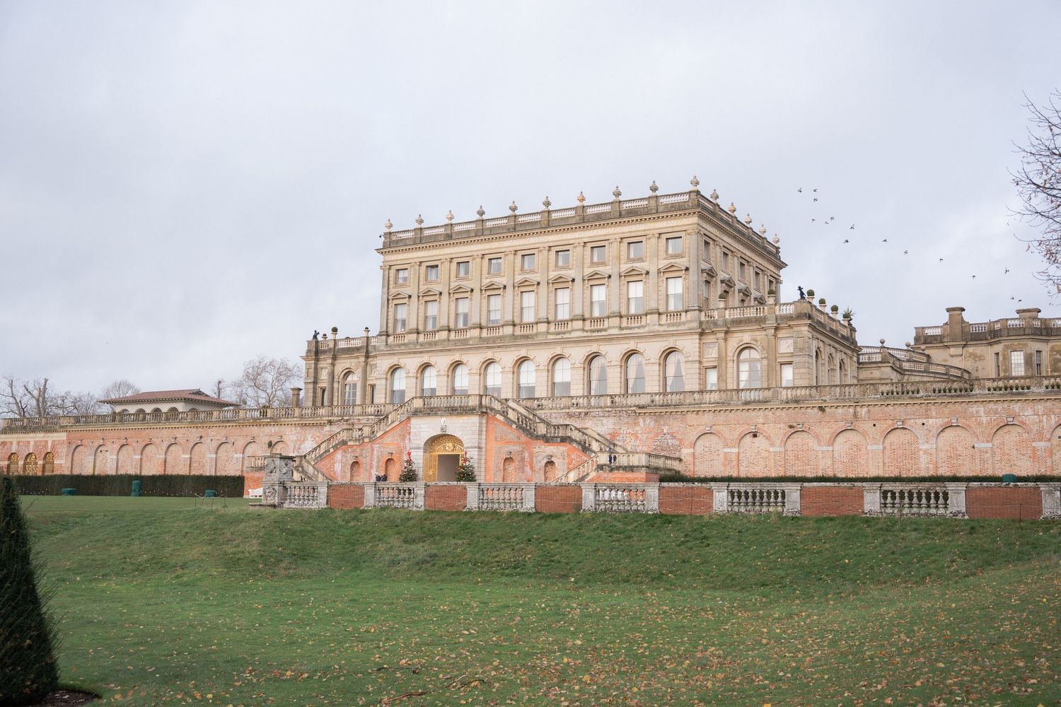 Grand baroque palace with red brick facade, ornate stone balustrades, and manicured lawns under overcast sky.