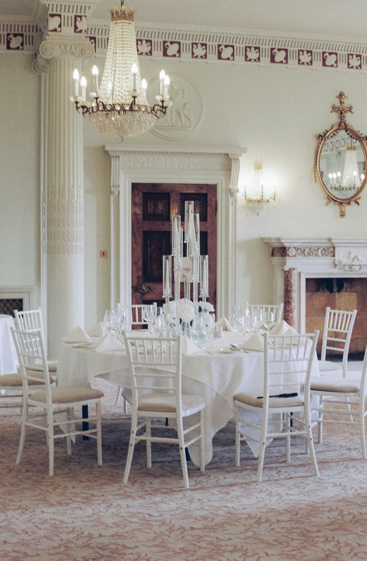 Elegant white wedding reception setup with chandelier and white chiavari chairs in a grand room with ornate details.