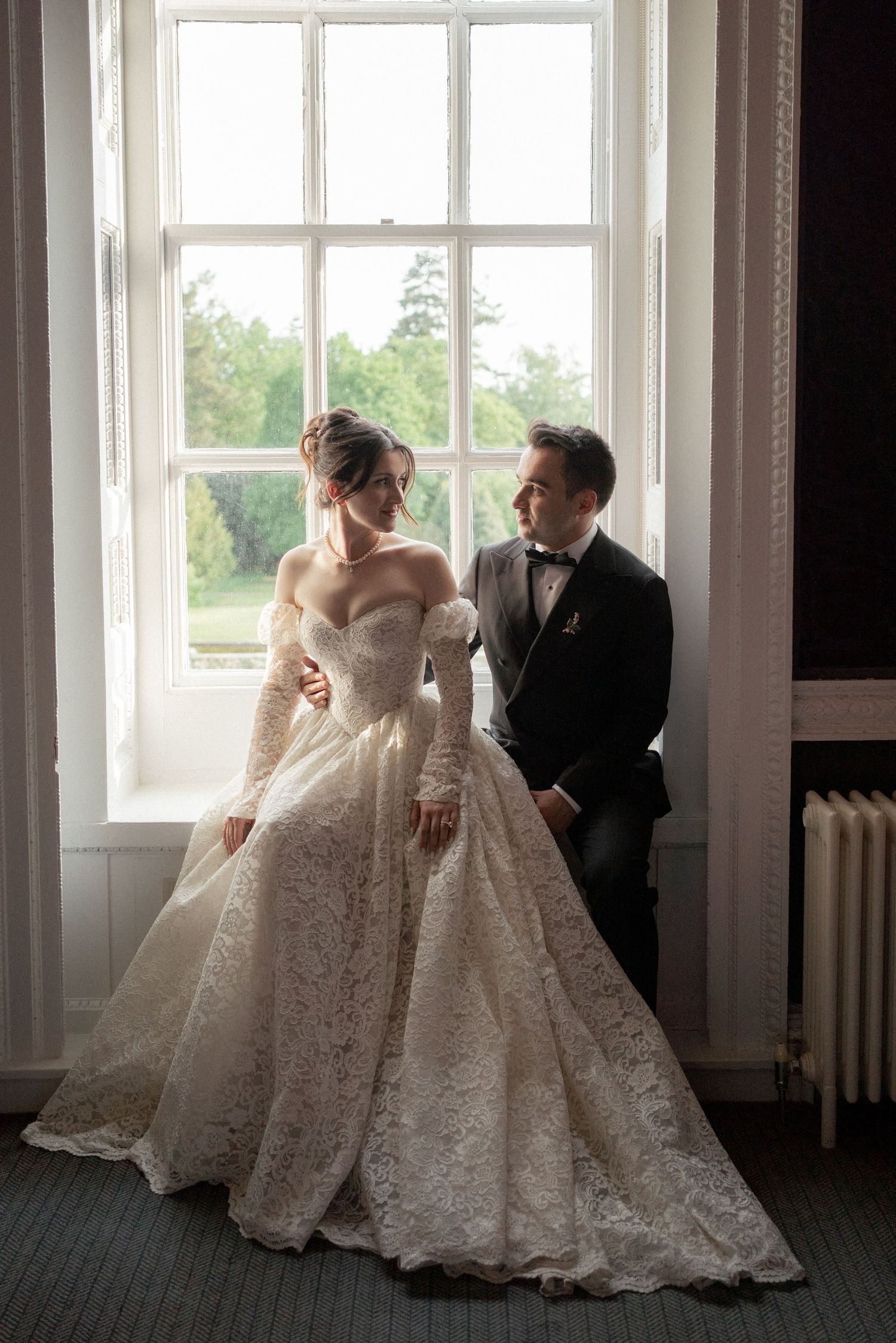 A romantic wedding moment by a bright window featuring an elegant lace gown and classic black tuxedo.