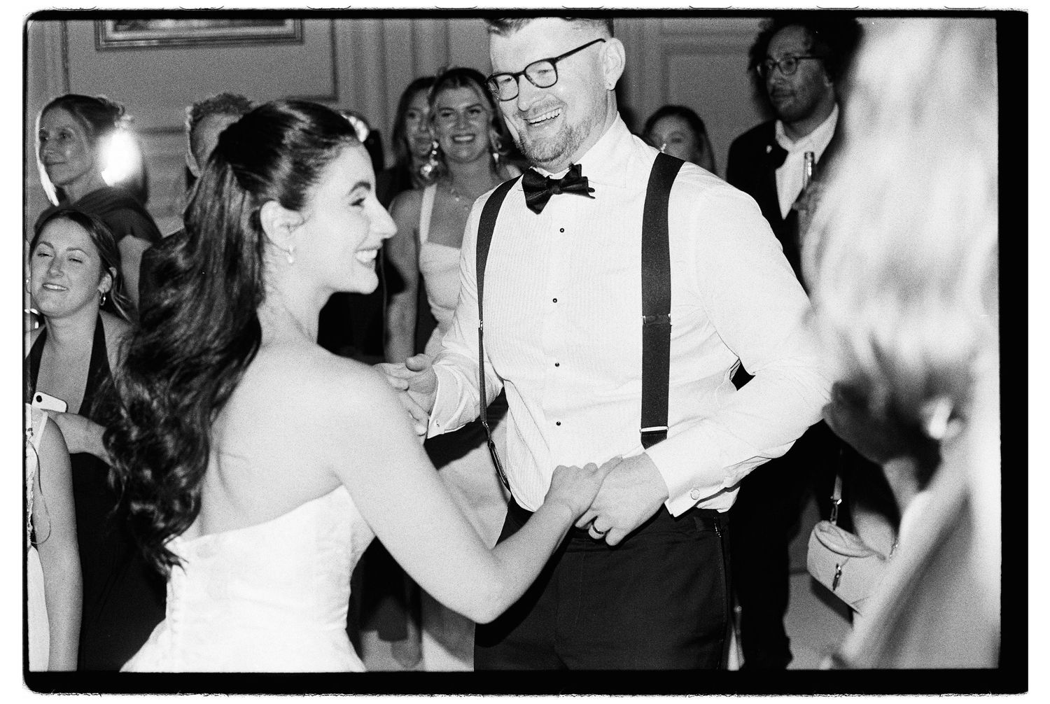 Bride and groom share first dance surrounded by smiling guests at black and white film wedding reception.