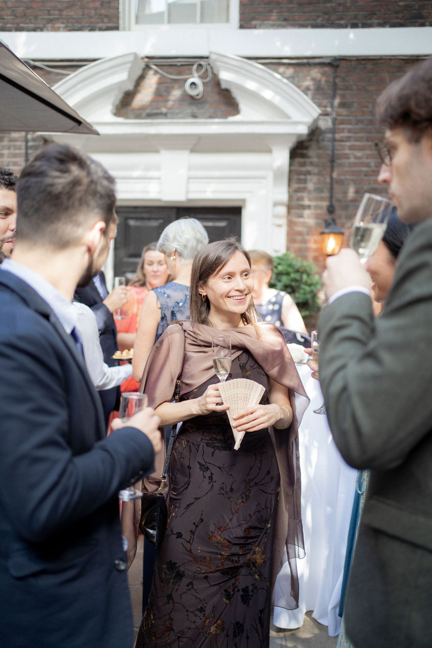 A smiling woman in a dark floral gown holds a fan at an elegant outdoor garden party with guests in formal attire.