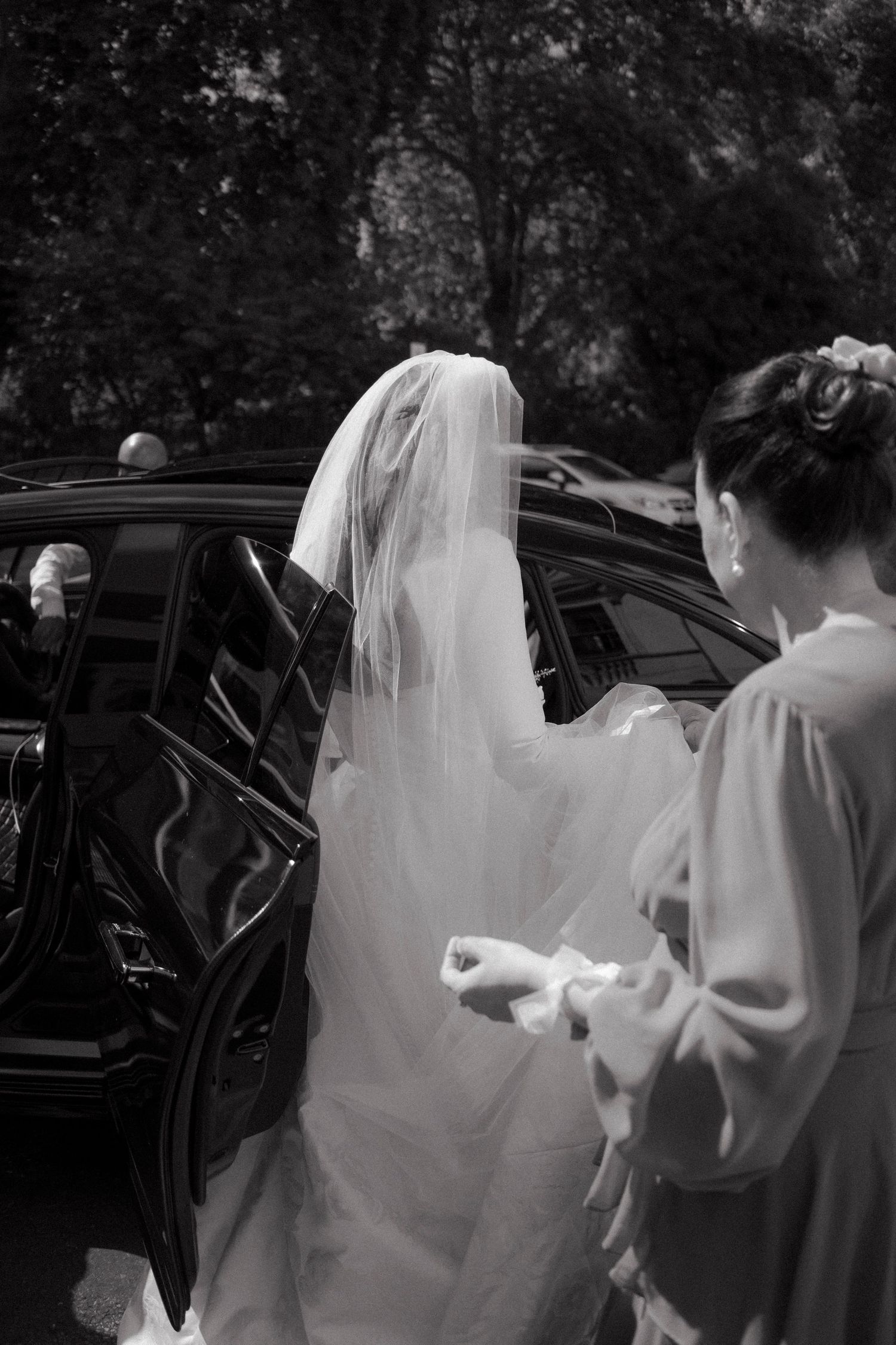 Black and white photo of a bride in white gown and veil getting into a car, assisted by a bridesmaid.