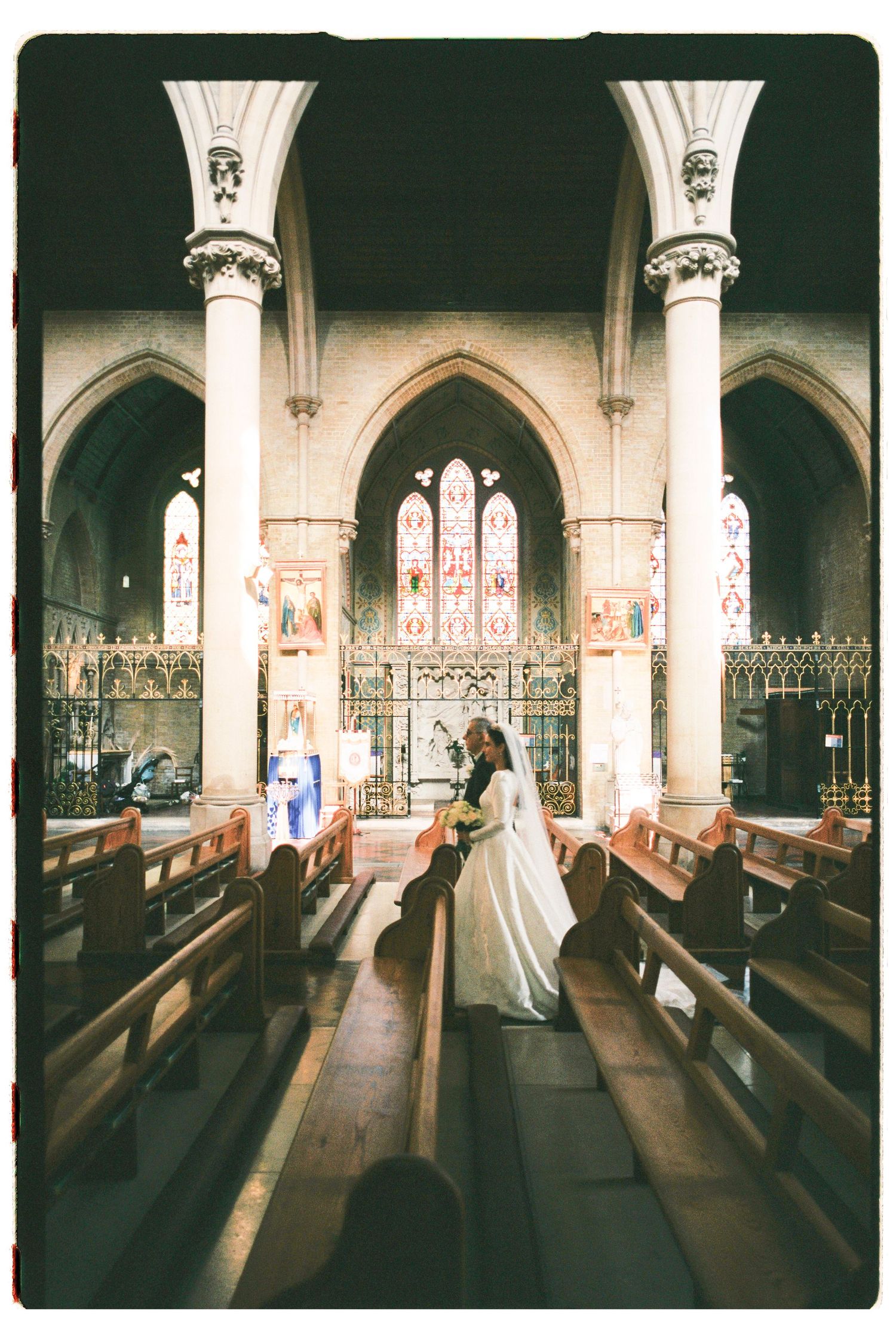 Bride and groom share a romantic moment in a stunning Gothic church aisle, surrounded by ornate stained glass windows and wooden pews.