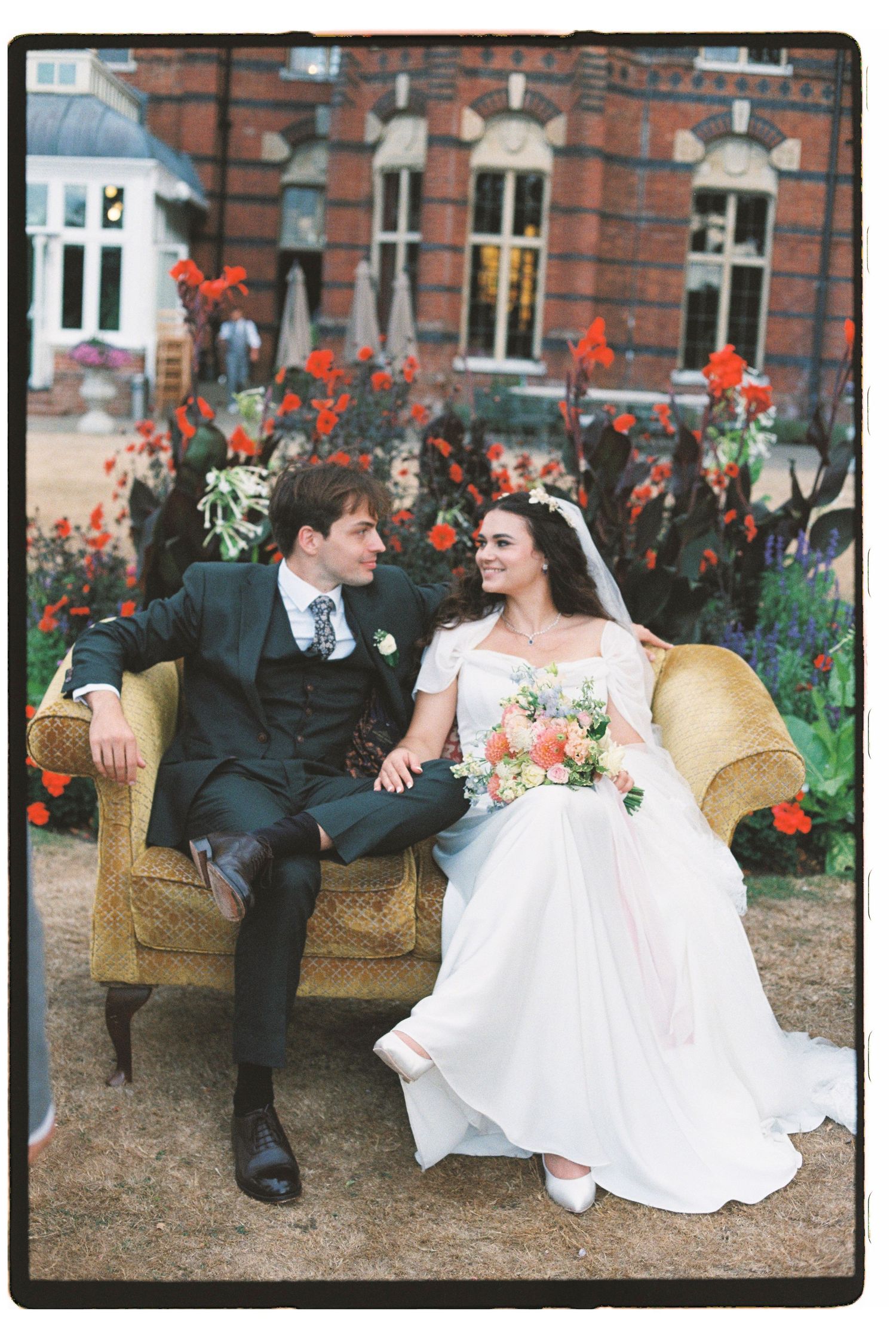 Newlywed couple sits on vintage sofa in front of red brick manor with flowers.