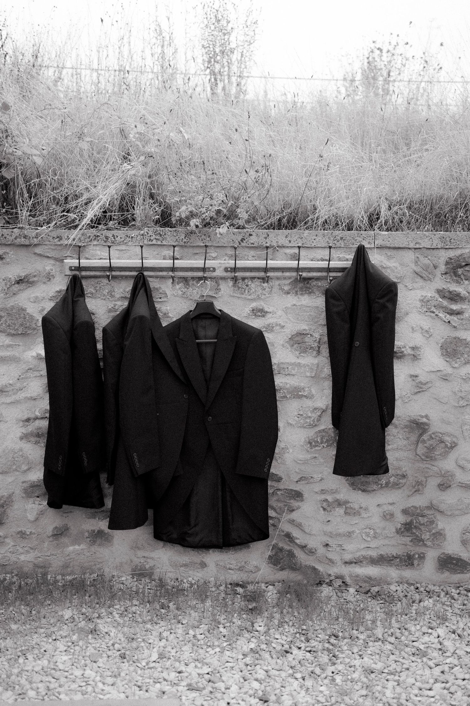 Three dark suit jackets hanging on hooks against rustic stone wall outdoors at wedding venue.