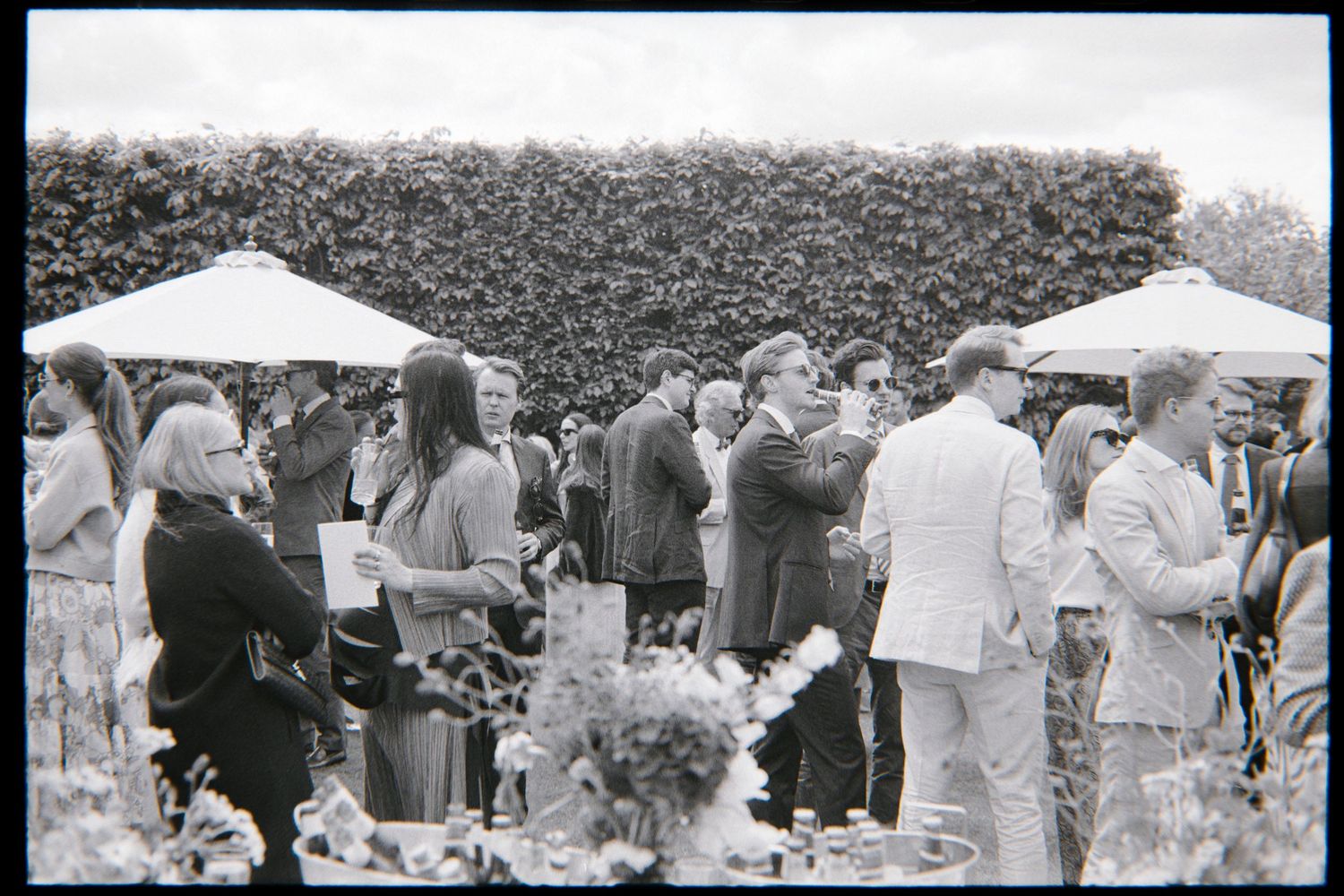 Wedding guests in summer attire chat and hold drinks beneath white parasols near a tall green hedge at an outdoor reception.