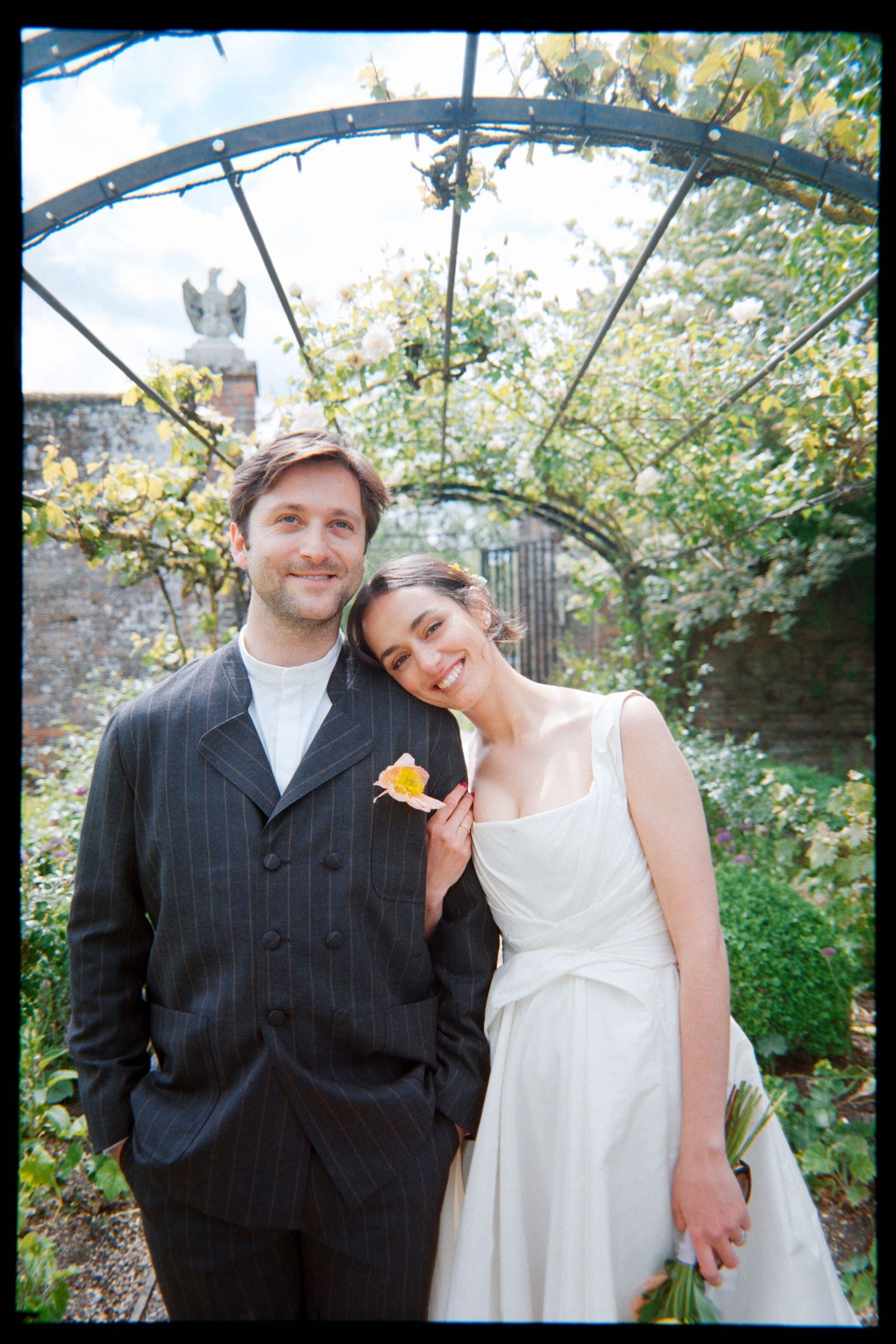 Smiling couple in a garden, the bride in a white dress leaning on a groom in a pinstripe suit under a metal arbor.