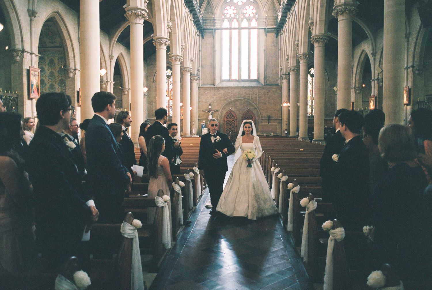 Bride in a white gown walks down the church aisle on a man's arm as wedding guests stand beside pews decorated with white bows.