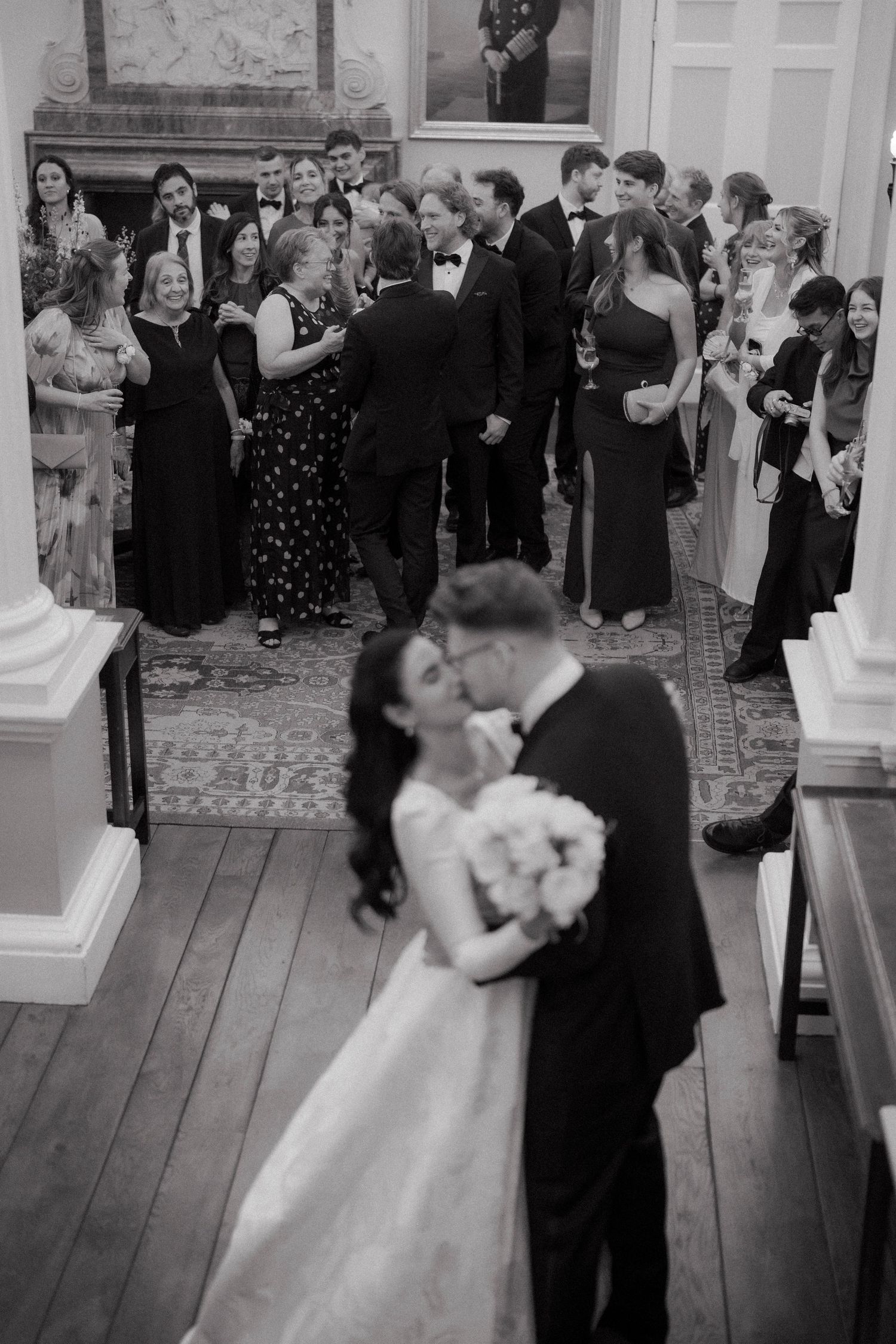 Black and white photo of bride and groom kissing with wedding guests mingling in background.