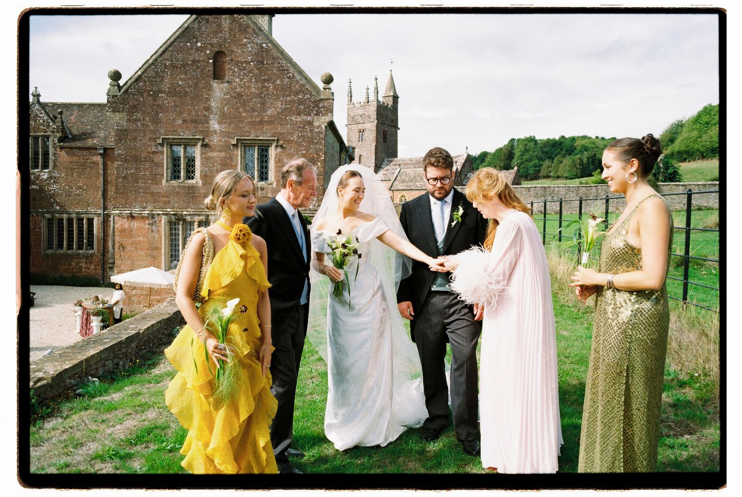 Bride and groom with wedding party in colorful dresses outside historic stone church after ceremony.