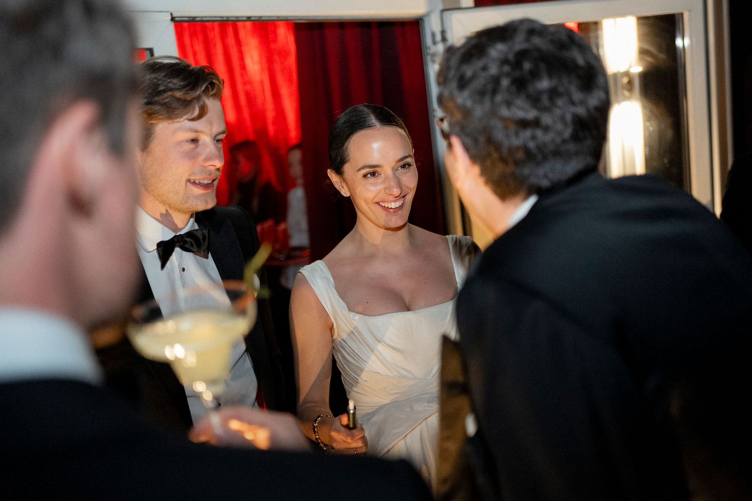 A smiling woman in a white gown chats with a man in a bow tie and tuxedo at an elegant evening party with red drapes.