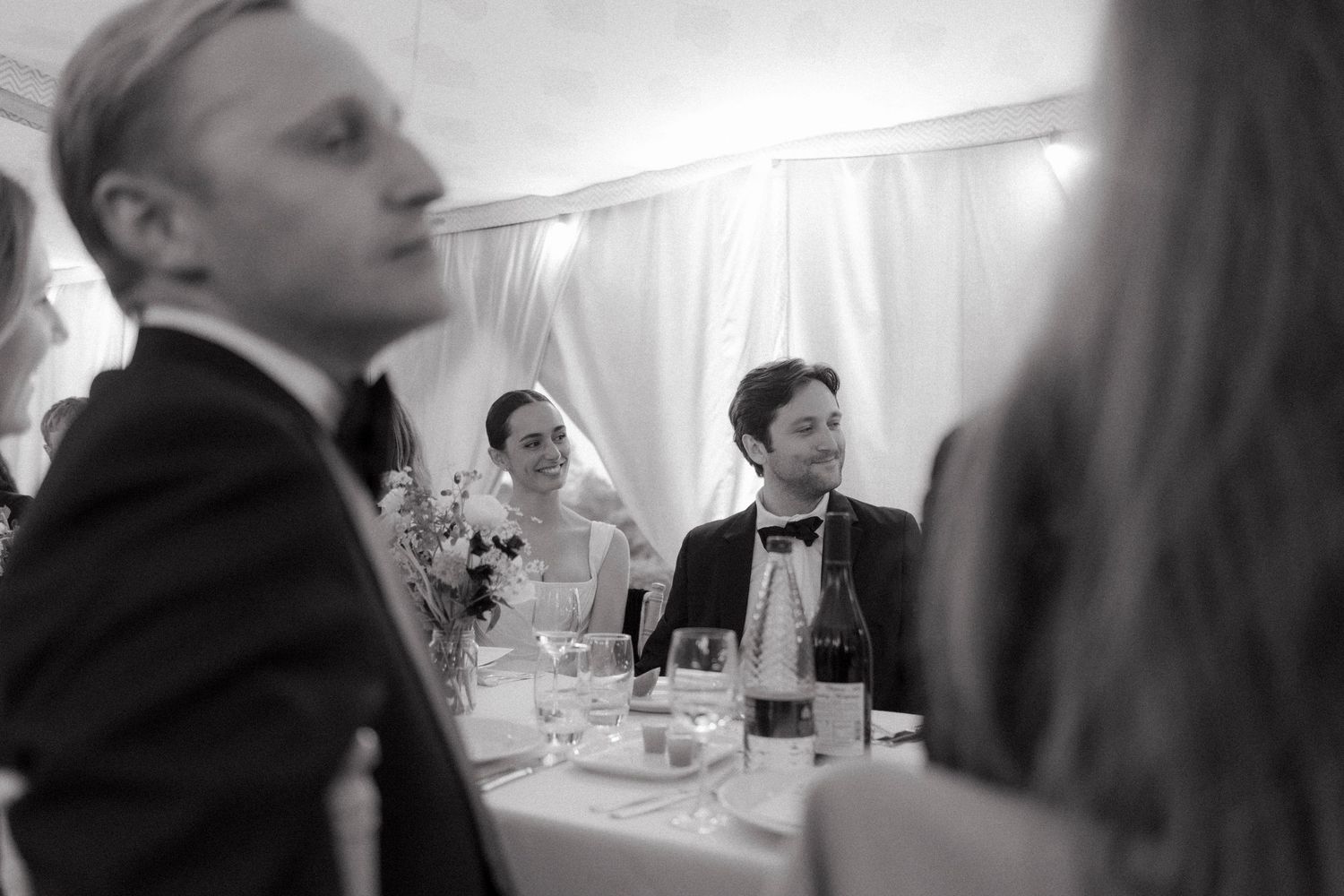 Black and white photo of wedding guests in formal attire seated at a reception table with glasses and flowers, smiling and looking on.
