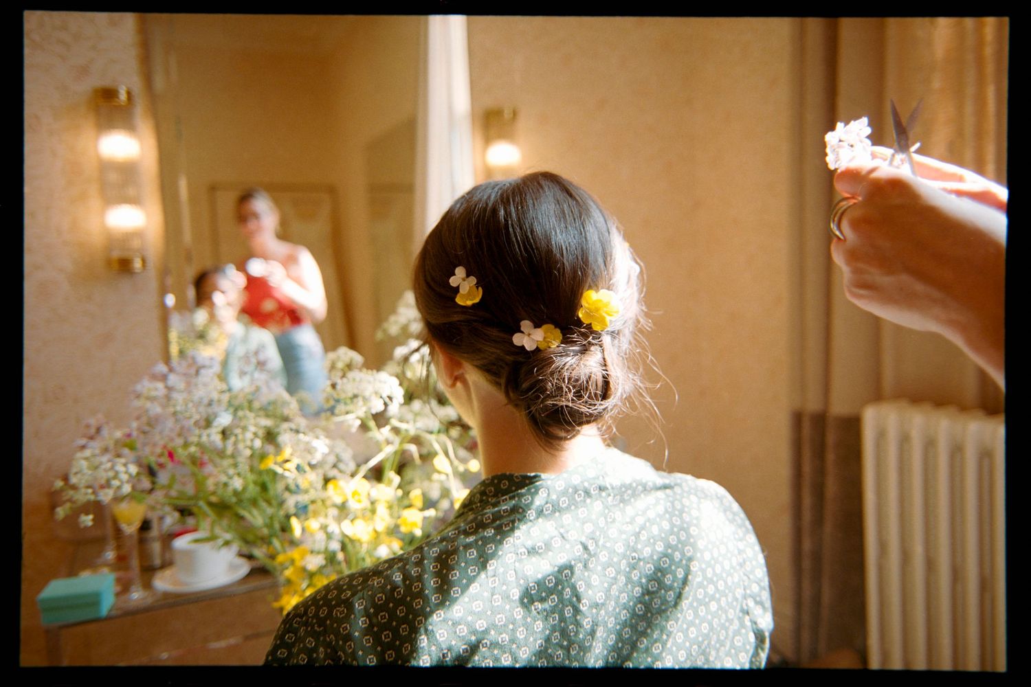 Woman seen from behind with small yellow flowers pinned in her upswept hair, facing a mirror beside a bouquet of white and yellow blooms.