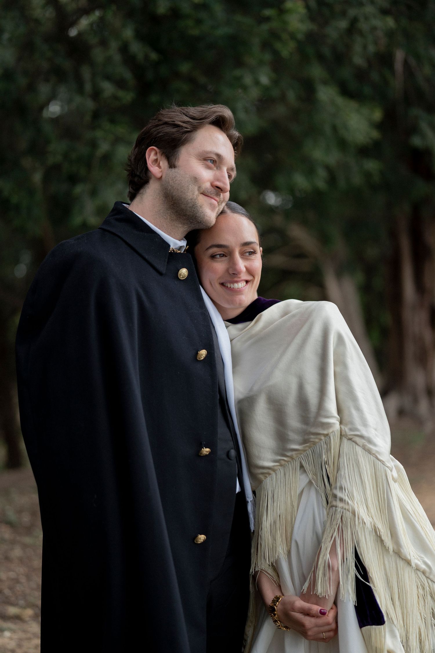 Happy couple standing close outdoors among tall trees, he in a navy cape coat and she in a cream fringed shawl.