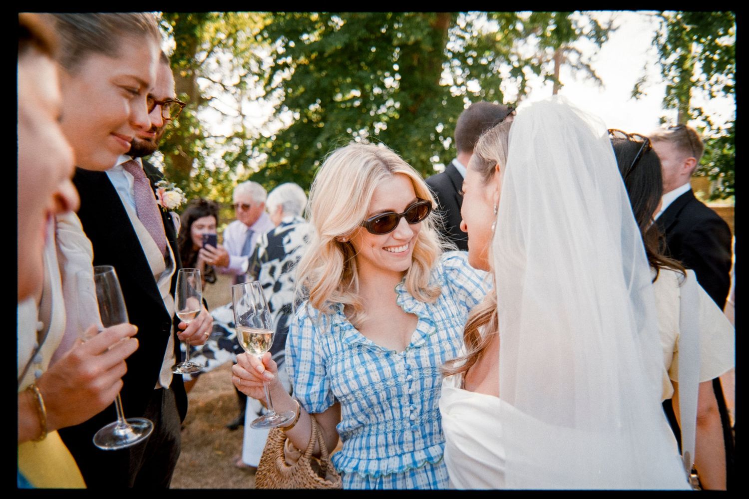 Smiling blonde woman in sunglasses and a blue plaid dress chats with a veiled bride among wedding guests at an outdoor garden reception.