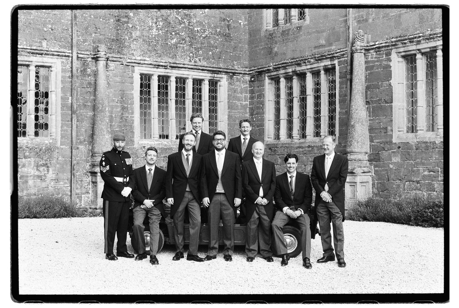 Black and white photo of nine men in suits and a uniformed officer posing together in front of a historic stone building.