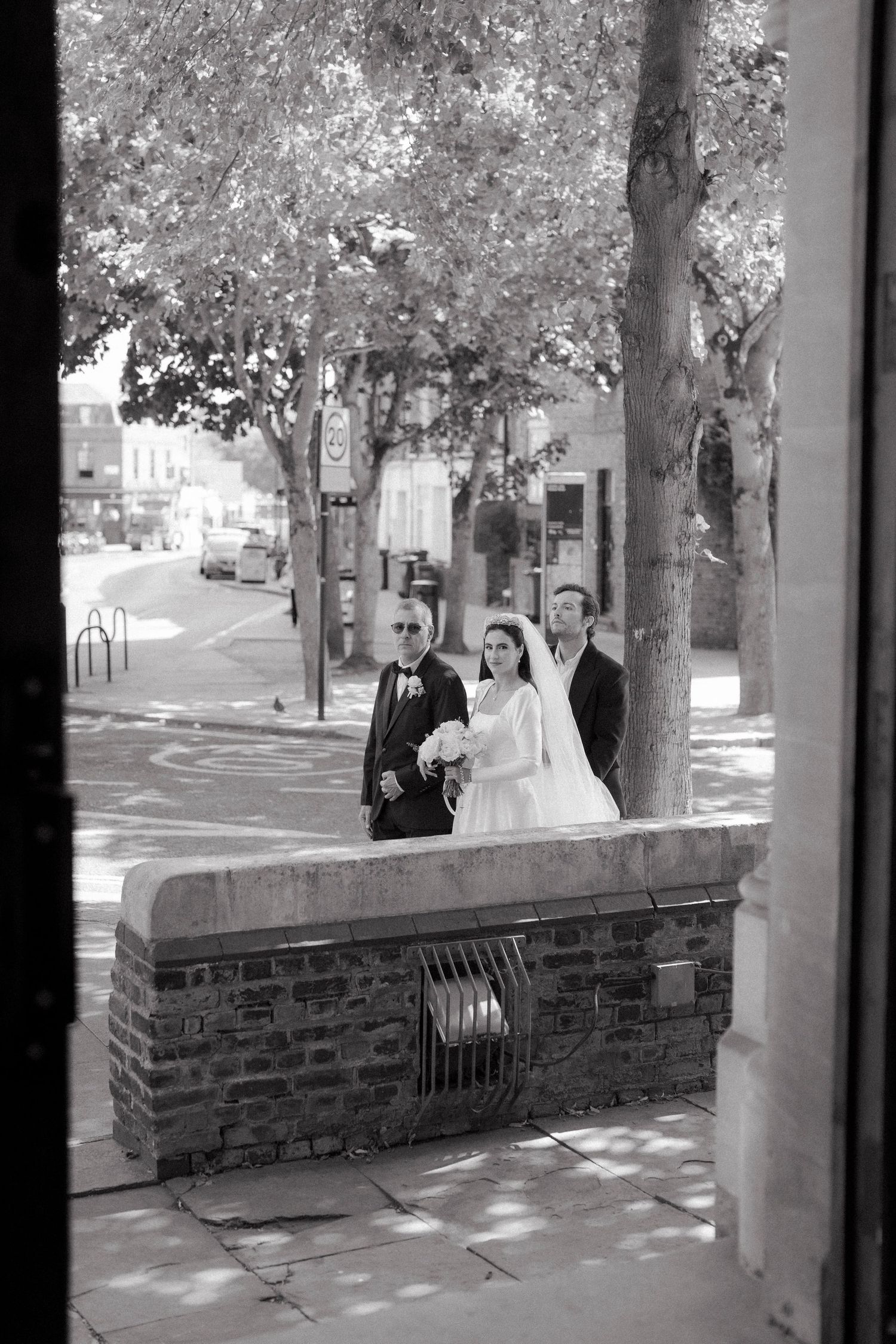 Black and white photo of a bride with two men in suits walking along a sunny sidewalk outside a church.