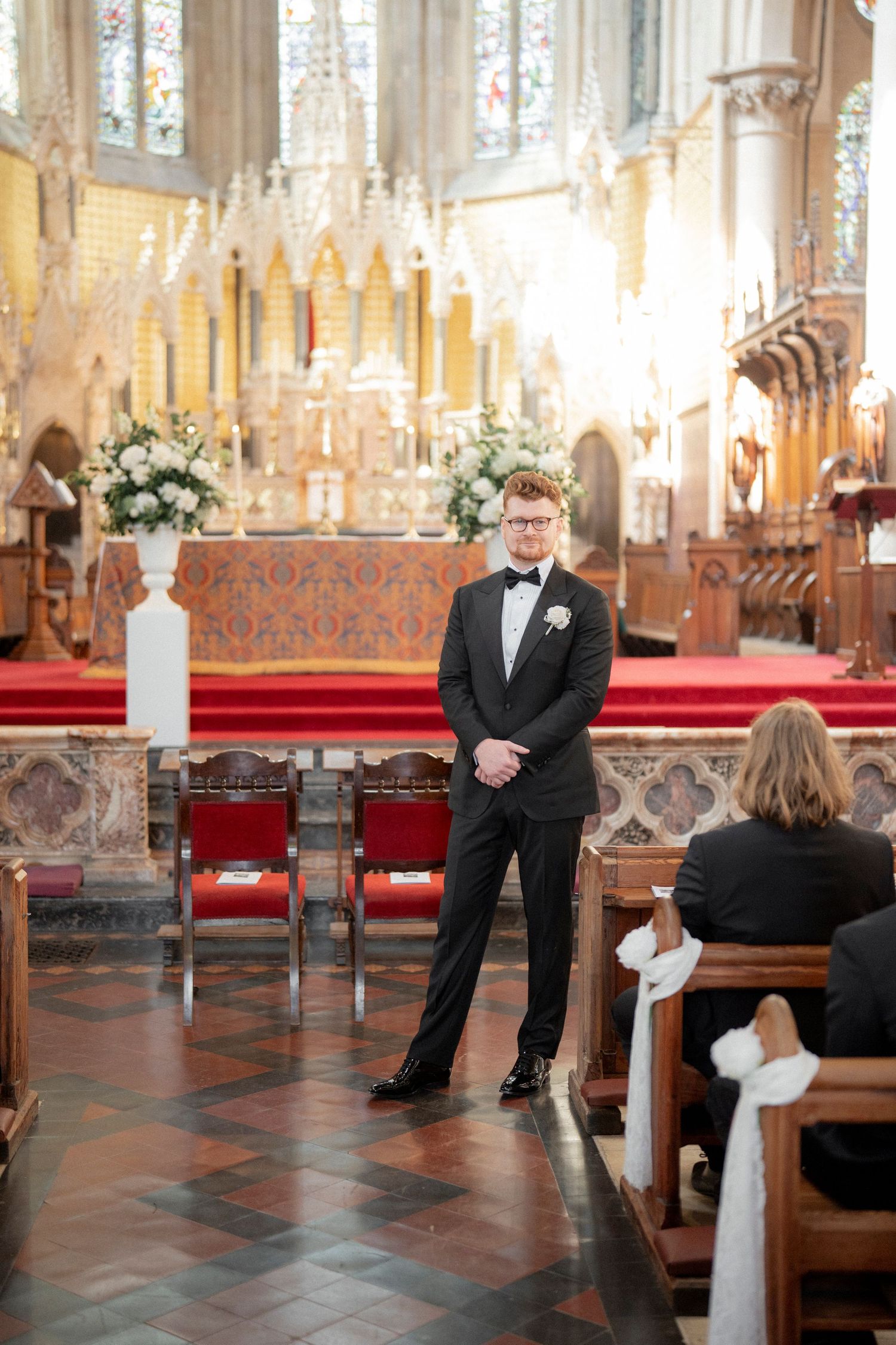 Groom in black tuxedo stands at church altar awaiting bride, with ornate red and gold decor behind him.