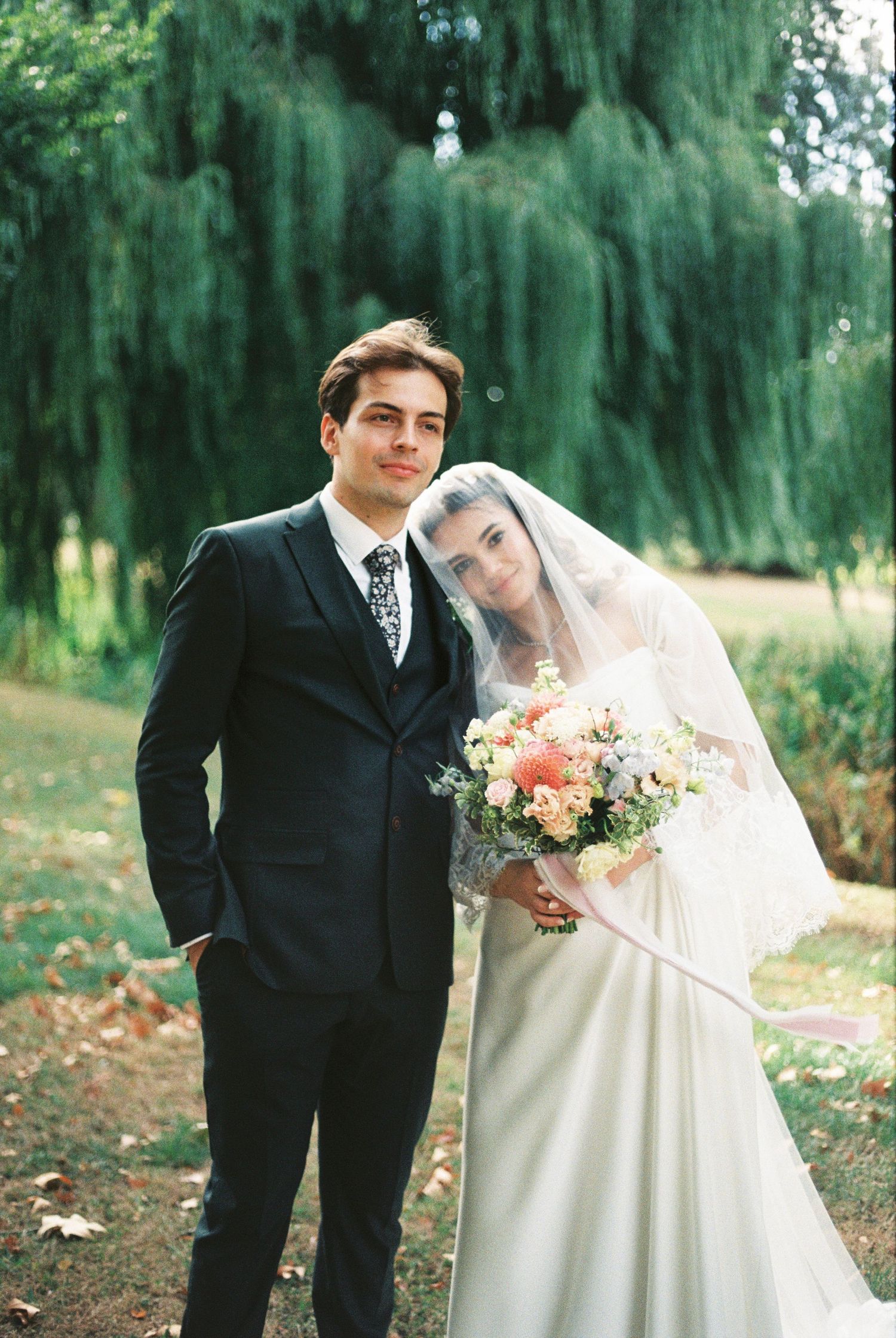 A young couple poses for a wedding portrait outdoors, bride in white gown with veil holding a bouquet, groom in dark suit.