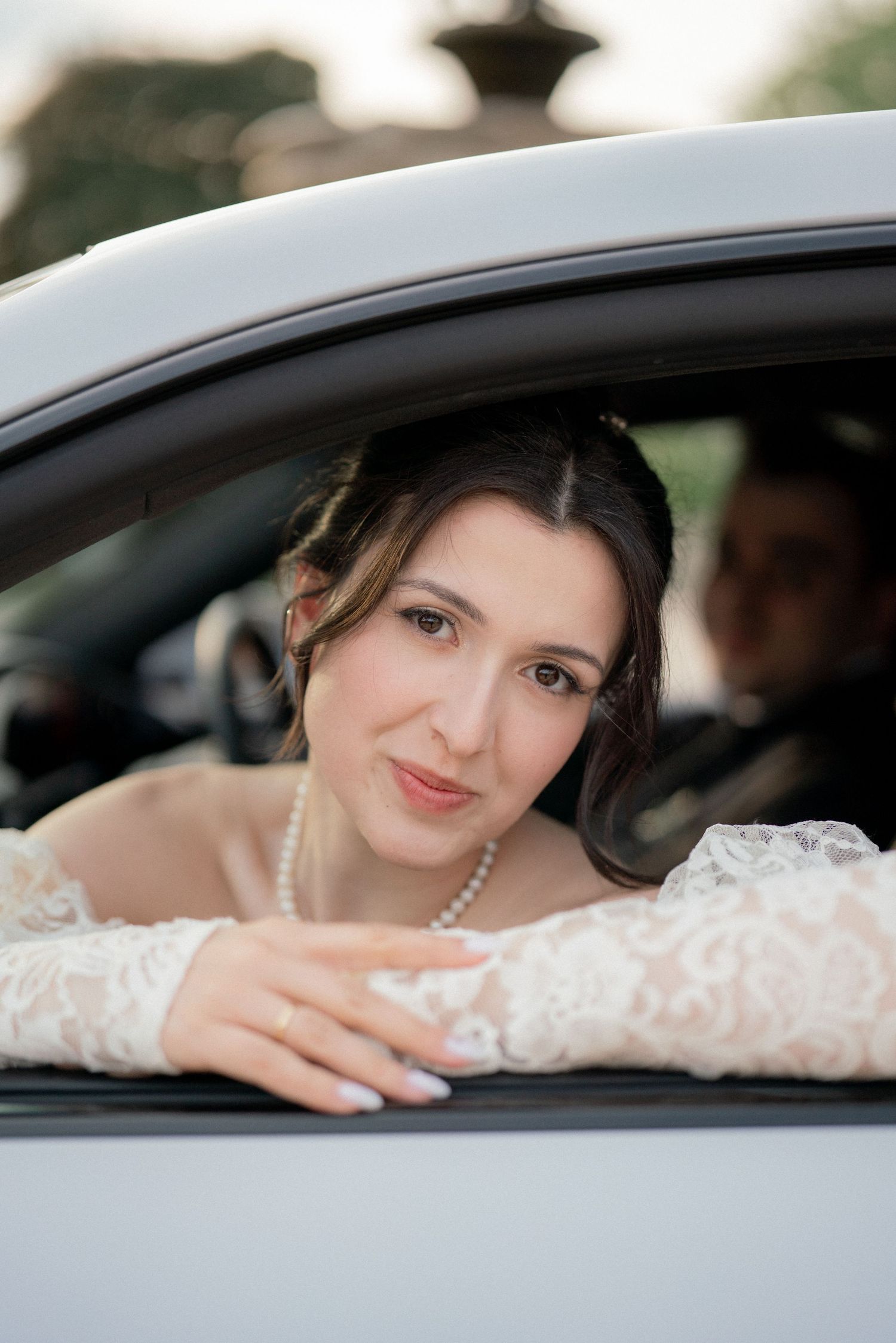 A close-up portrait through a car window showing delicate pearl jewelry and lace details.