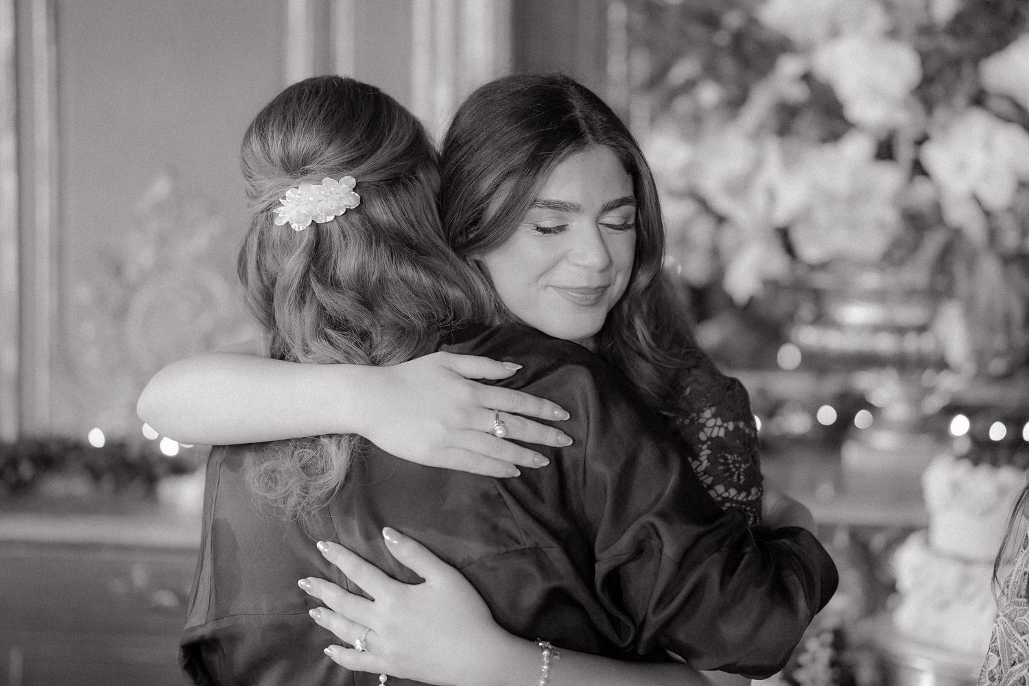 Two women share a warm embrace in a black and white photo, one wearing a floral hair accessory at an elegant venue.