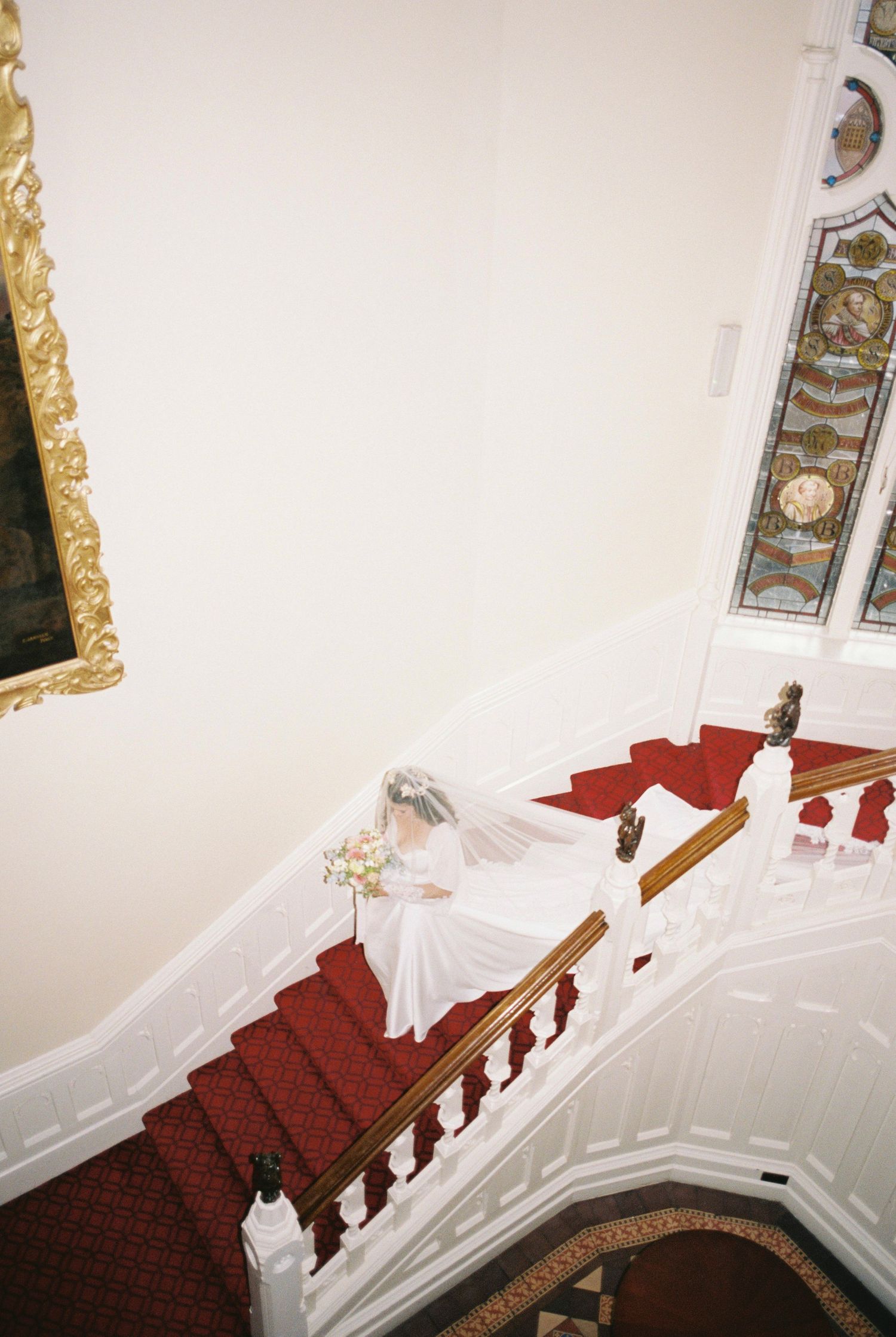 Bride in a white gown and veil holding a bouquet descends a red-carpeted staircase with a wooden banister beside a stained glass window.