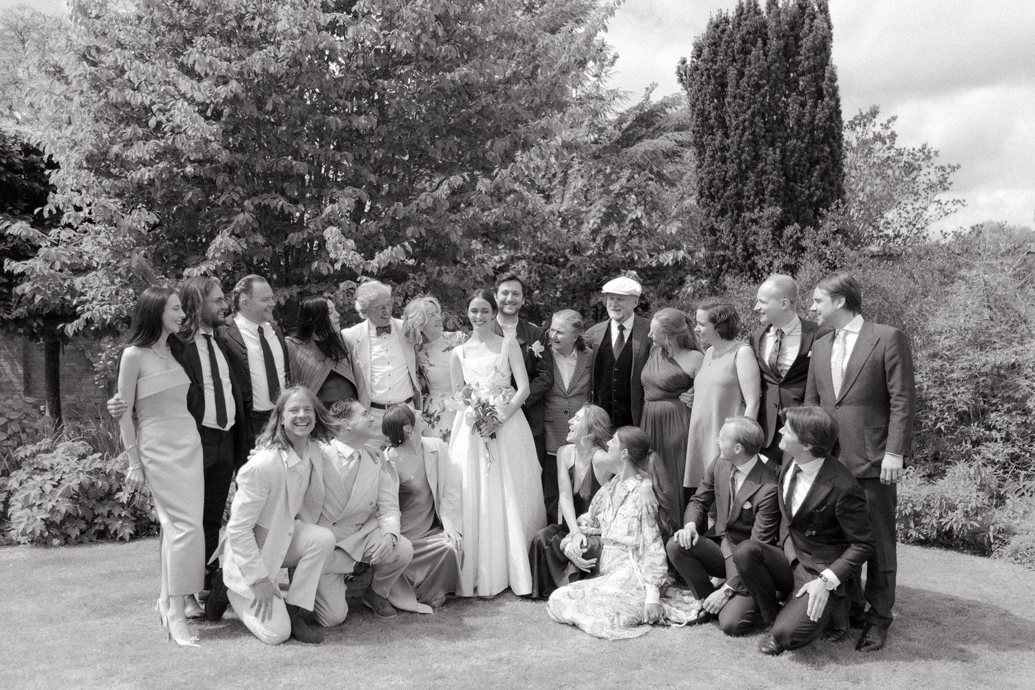 Black and white group portrait of a bride, groom and smiling wedding guests posed together on a lawn beside tall trees.
