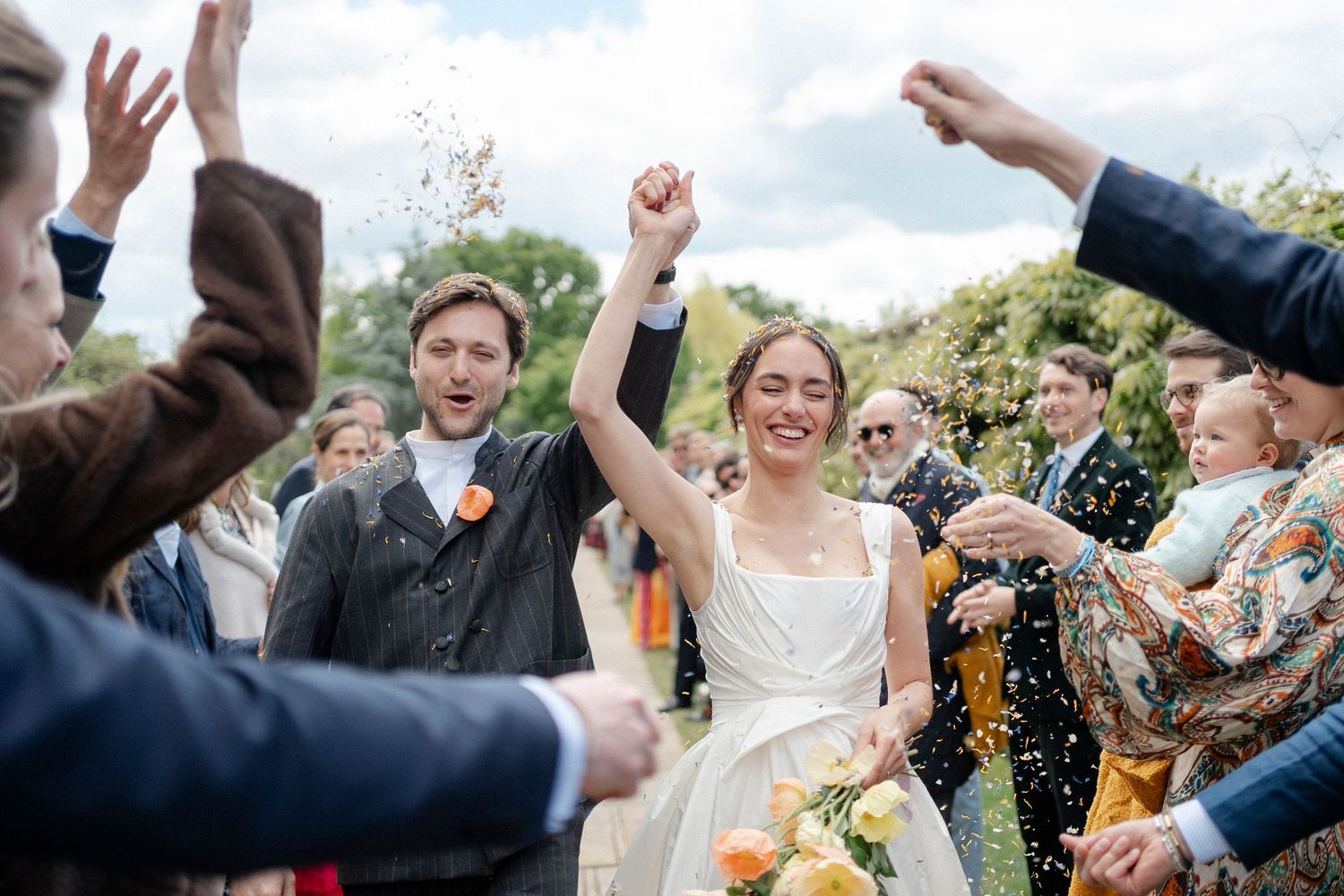 Joyful bride and groom raise their fists and smile as guests throw confetti at their outdoor wedding, the bride holding an orange bouquet.