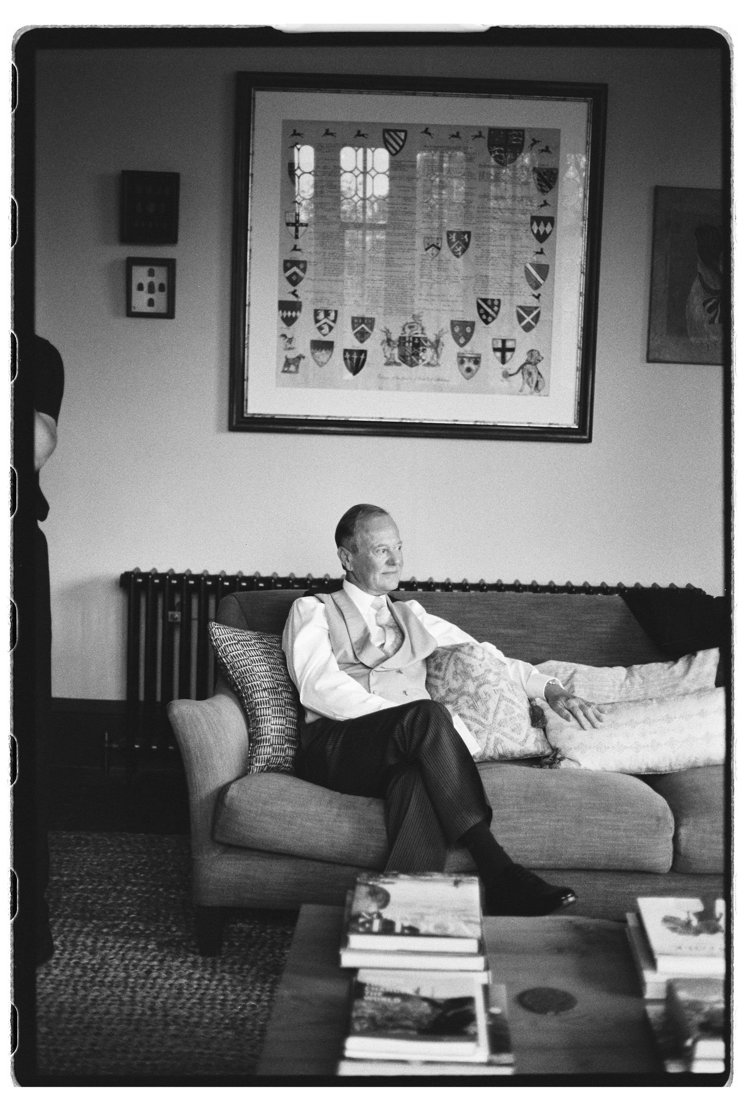 Black and white photo of an elderly man sitting on a sofa beneath a framed heraldic coat of arms artwork.