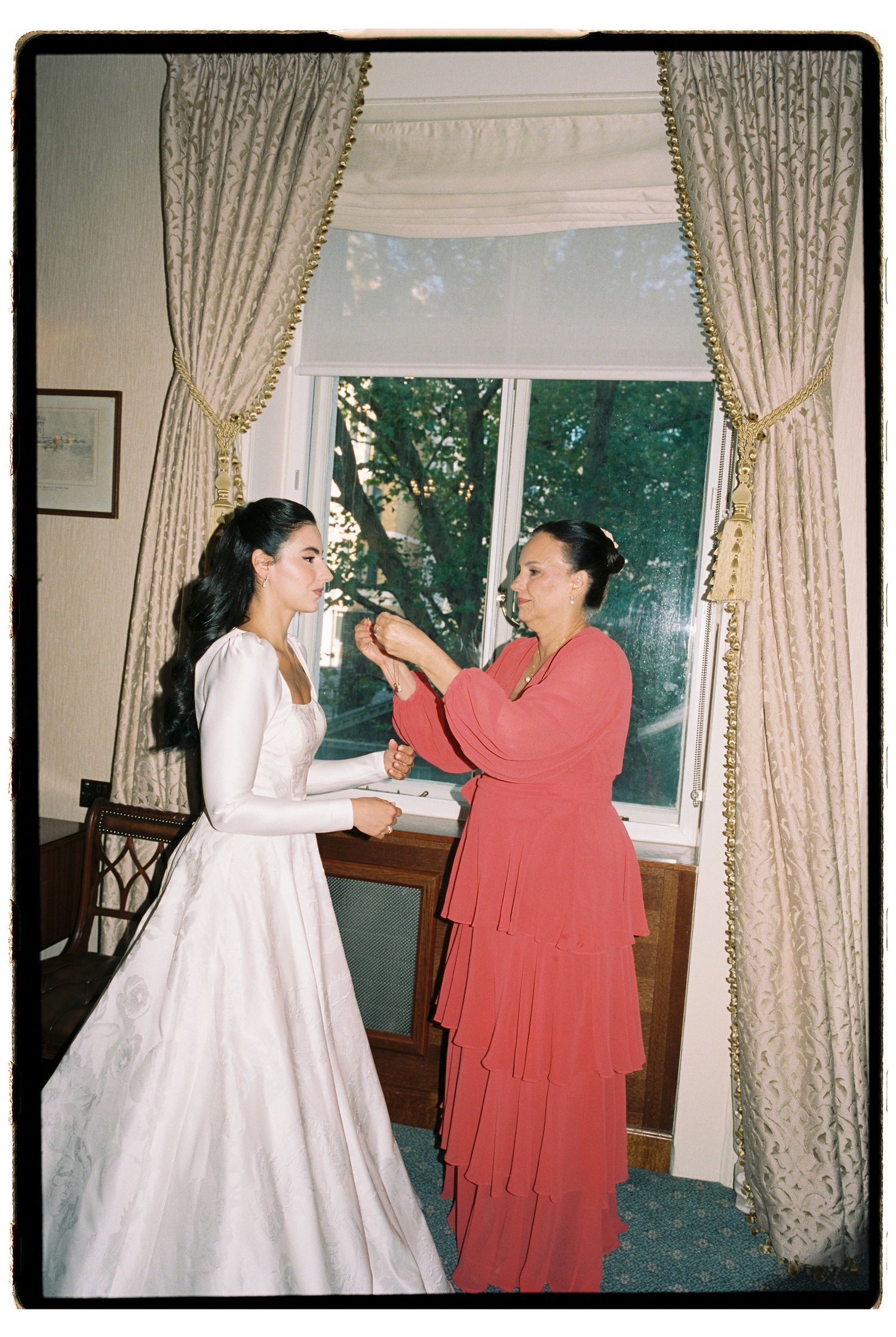 A bride in a white gown gets help from a woman in red near a draped window before her wedding ceremony.