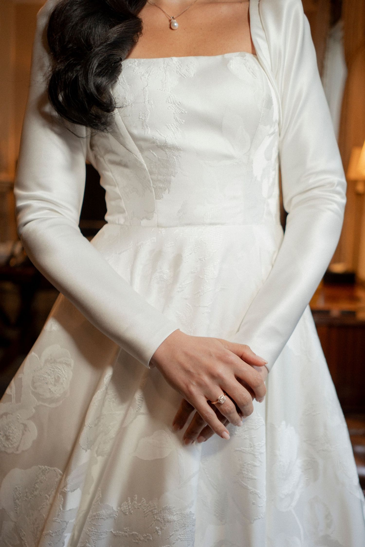 Bride in elegant long-sleeve white satin brocade wedding dress with floral detail, wearing a diamond ring.