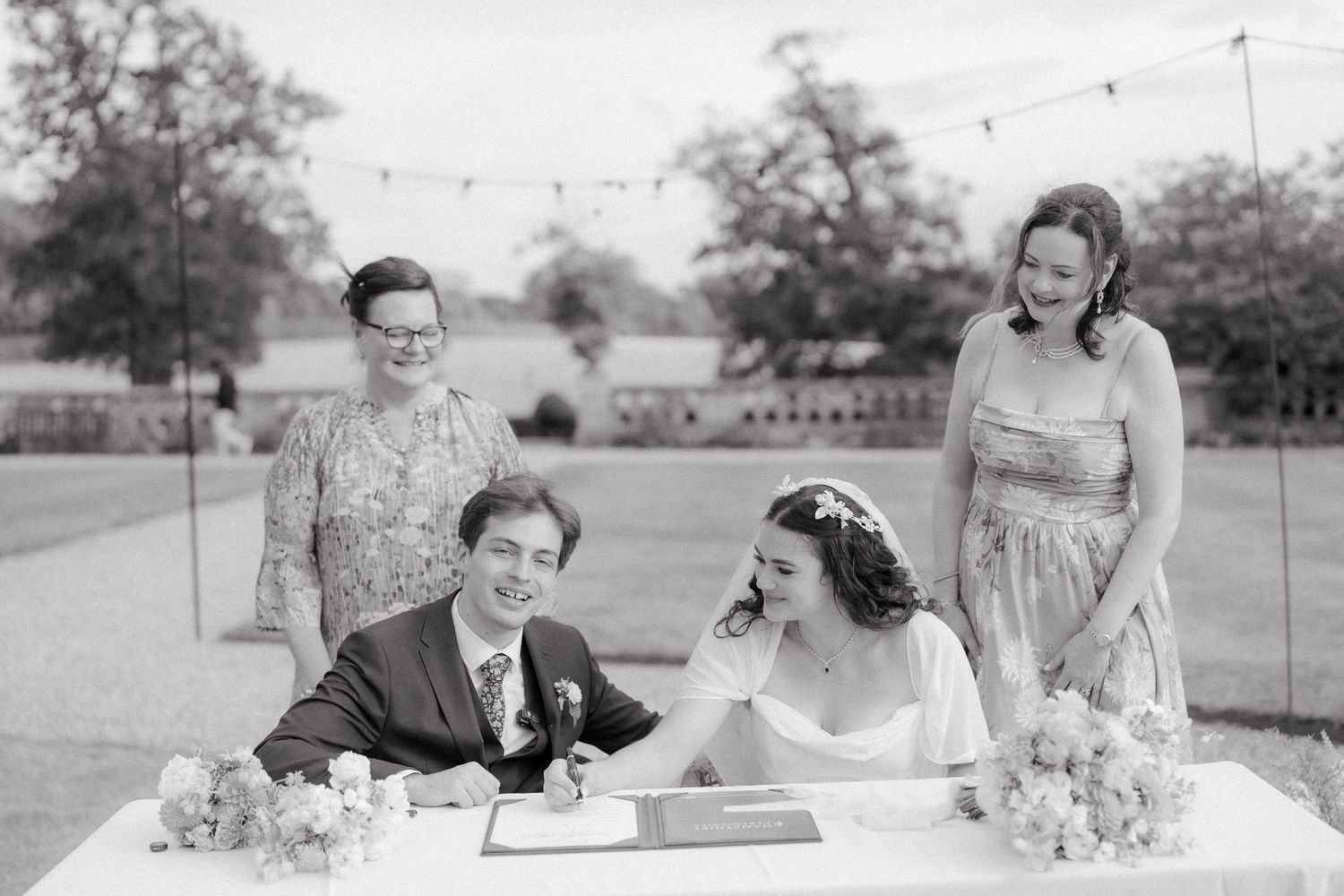 Black and white photo of a couple signing wedding register at outdoor ceremony with two witnesses smiling behind them.