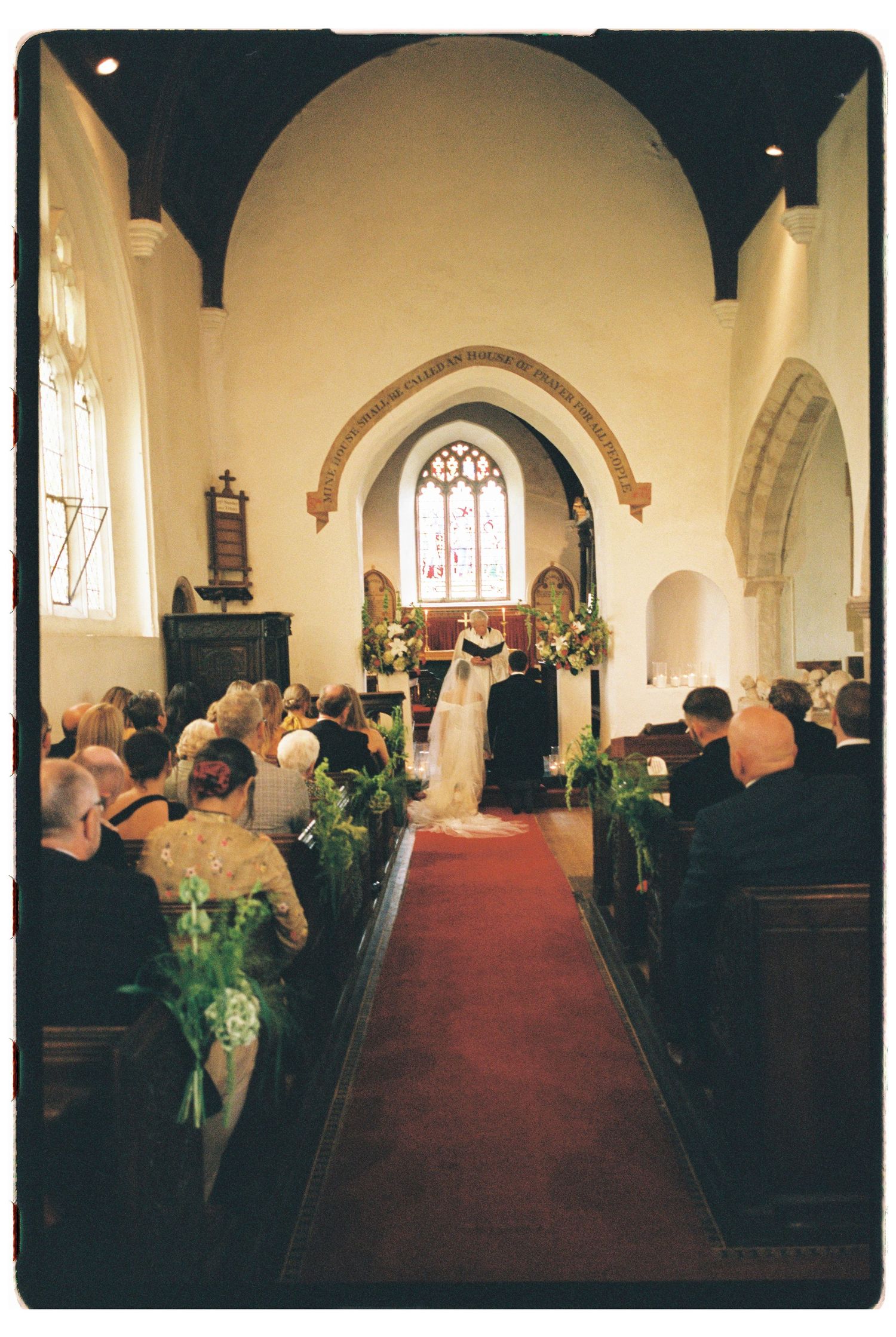 A bride and groom exchange vows at the altar of a charming traditional English church with red carpet aisle.
