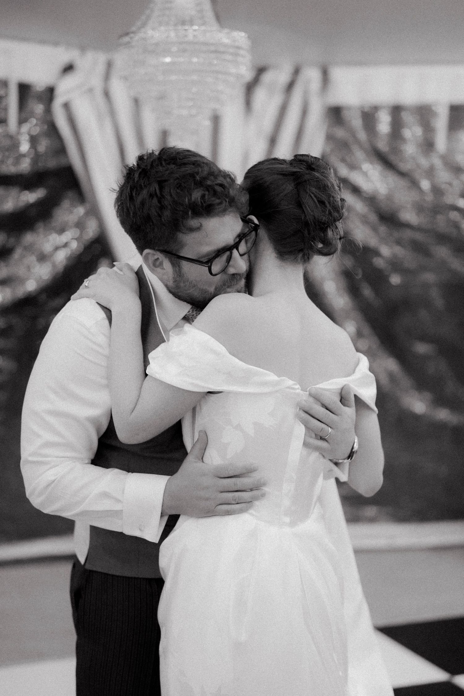 Newlyweds share intimate first dance embrace under chandelier at wedding reception.